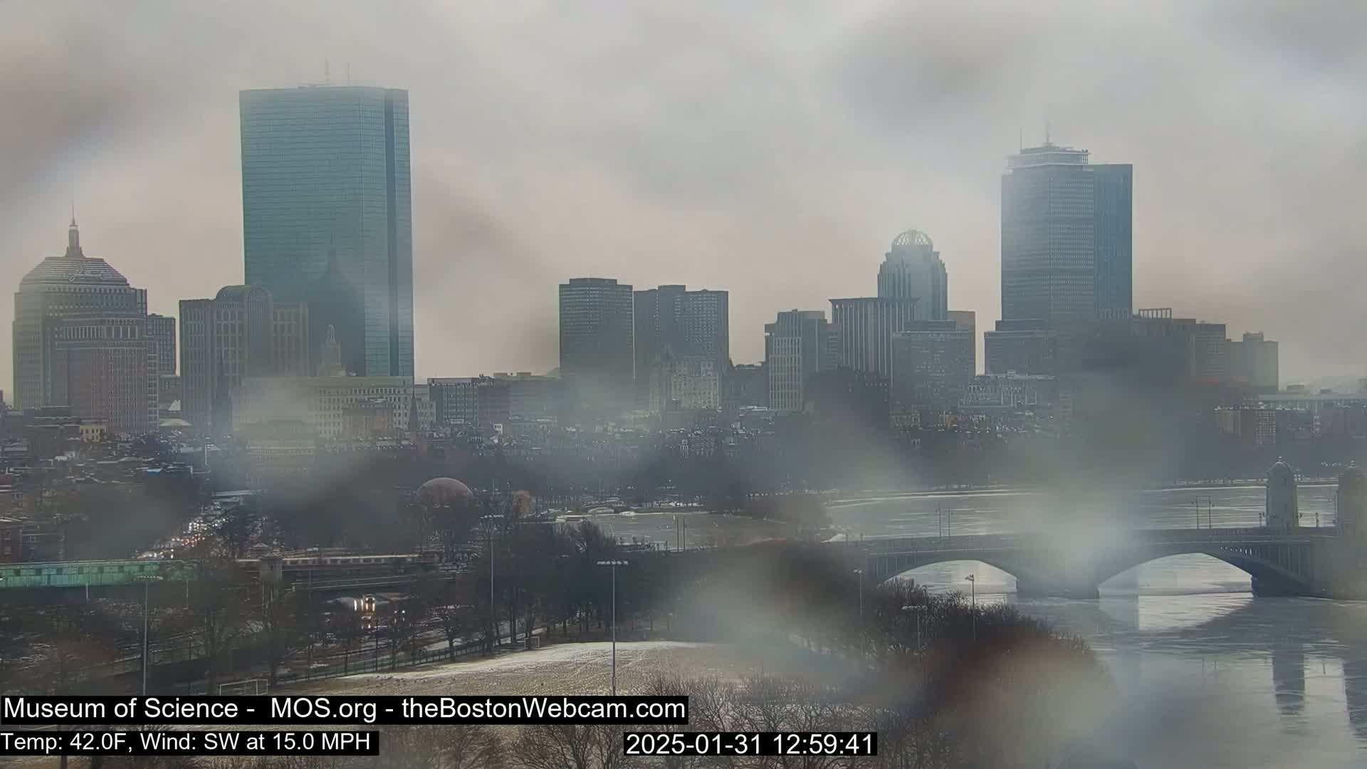 A hazy, snowy cityscape with a river and bridges is visible through a rain-streaked windowpane.