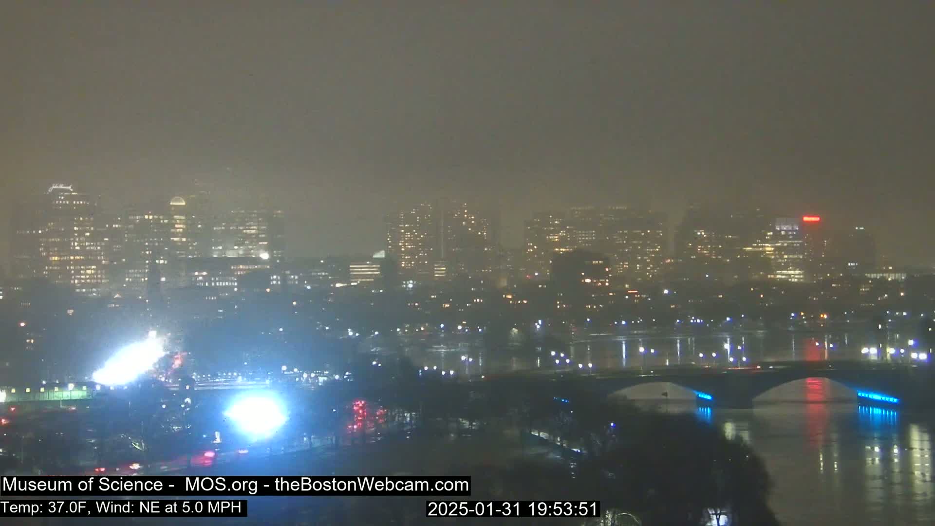 A foggy nighttime cityscape with a bridge illuminated over a body of water.