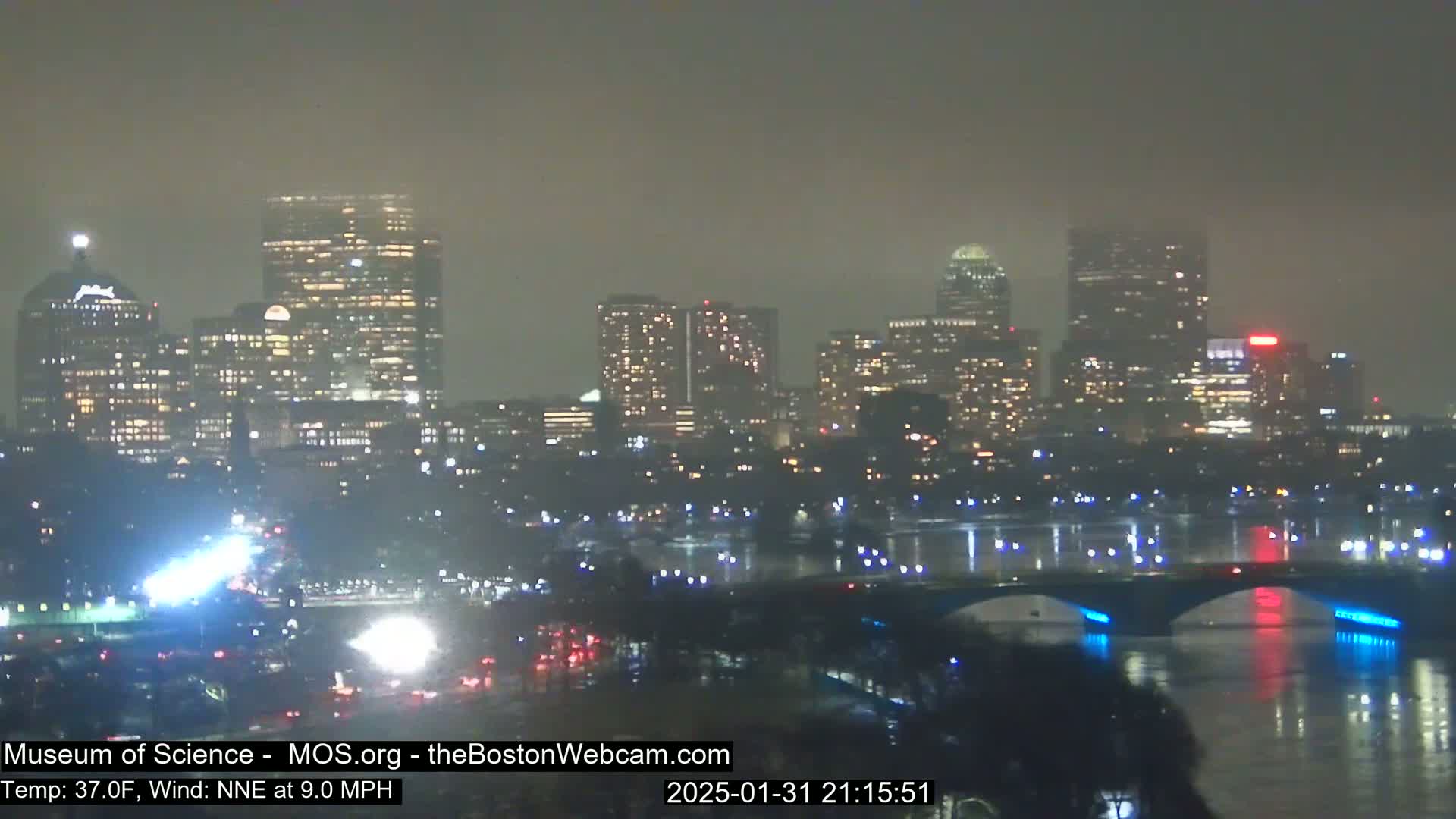 A hazy, nighttime view of a city skyline illuminated by lights, with a bridge and body of water in the foreground.