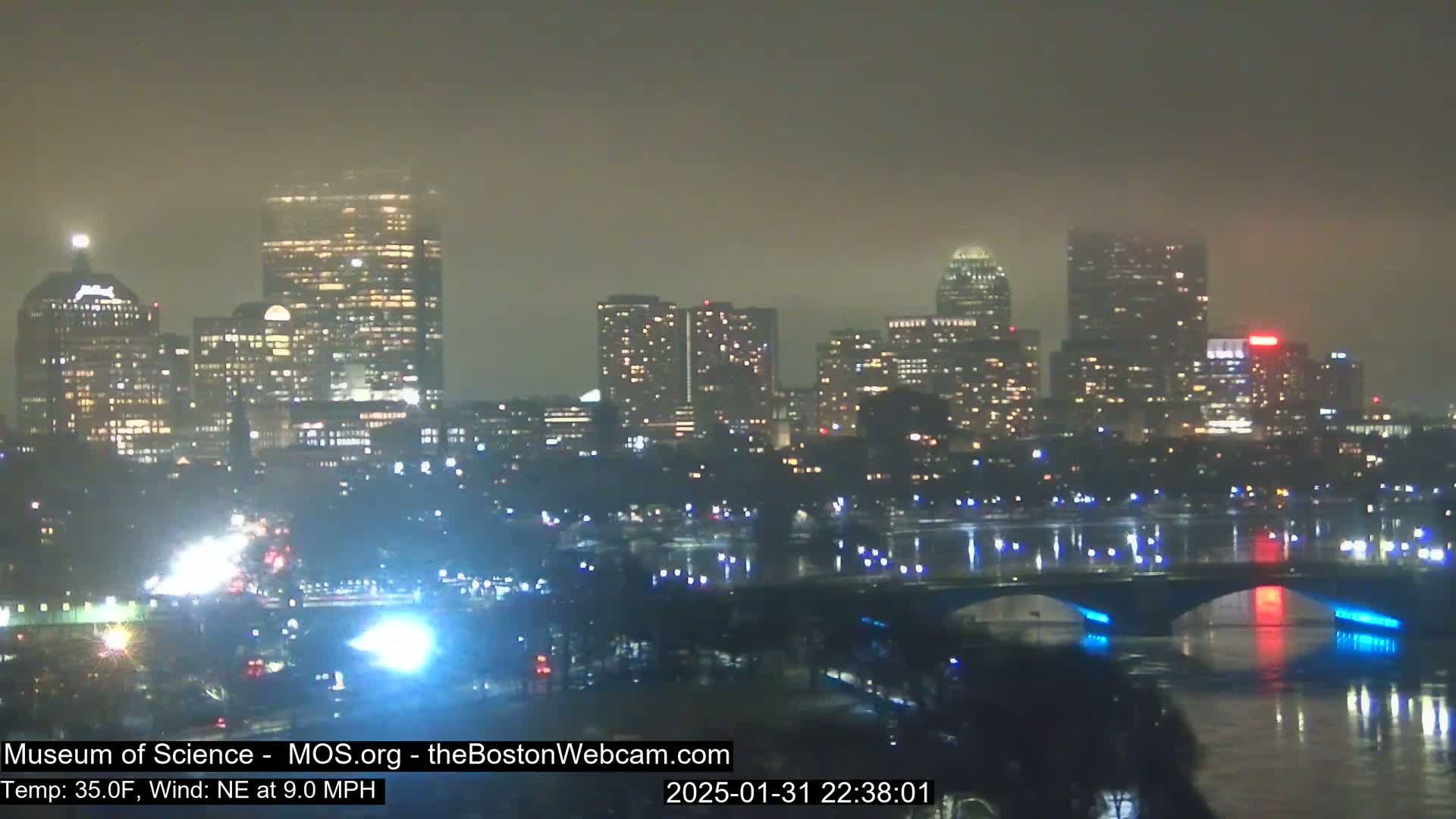 A nighttime cityscape with illuminated skyscrapers and bridges across a body of water under foggy conditions.