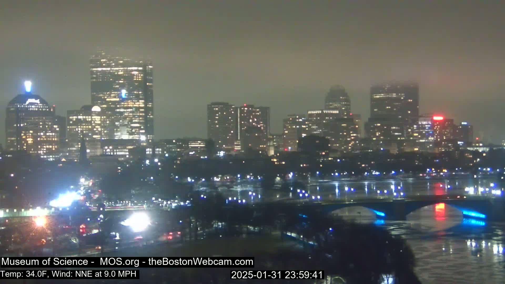 A nighttime, foggy view of a city skyline with illuminated buildings and a bridge over a river.