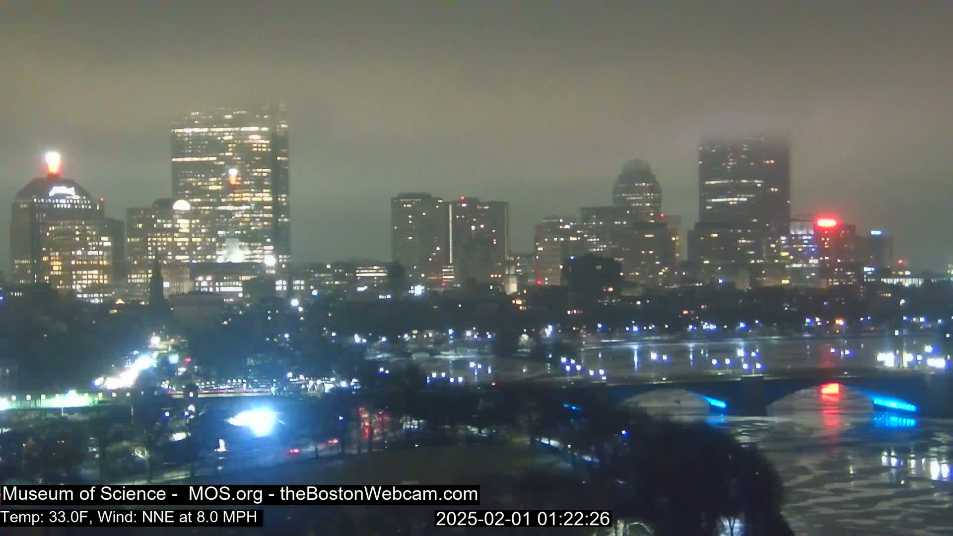 A foggy nighttime cityscape with illuminated buildings and a bridge over a body of water.