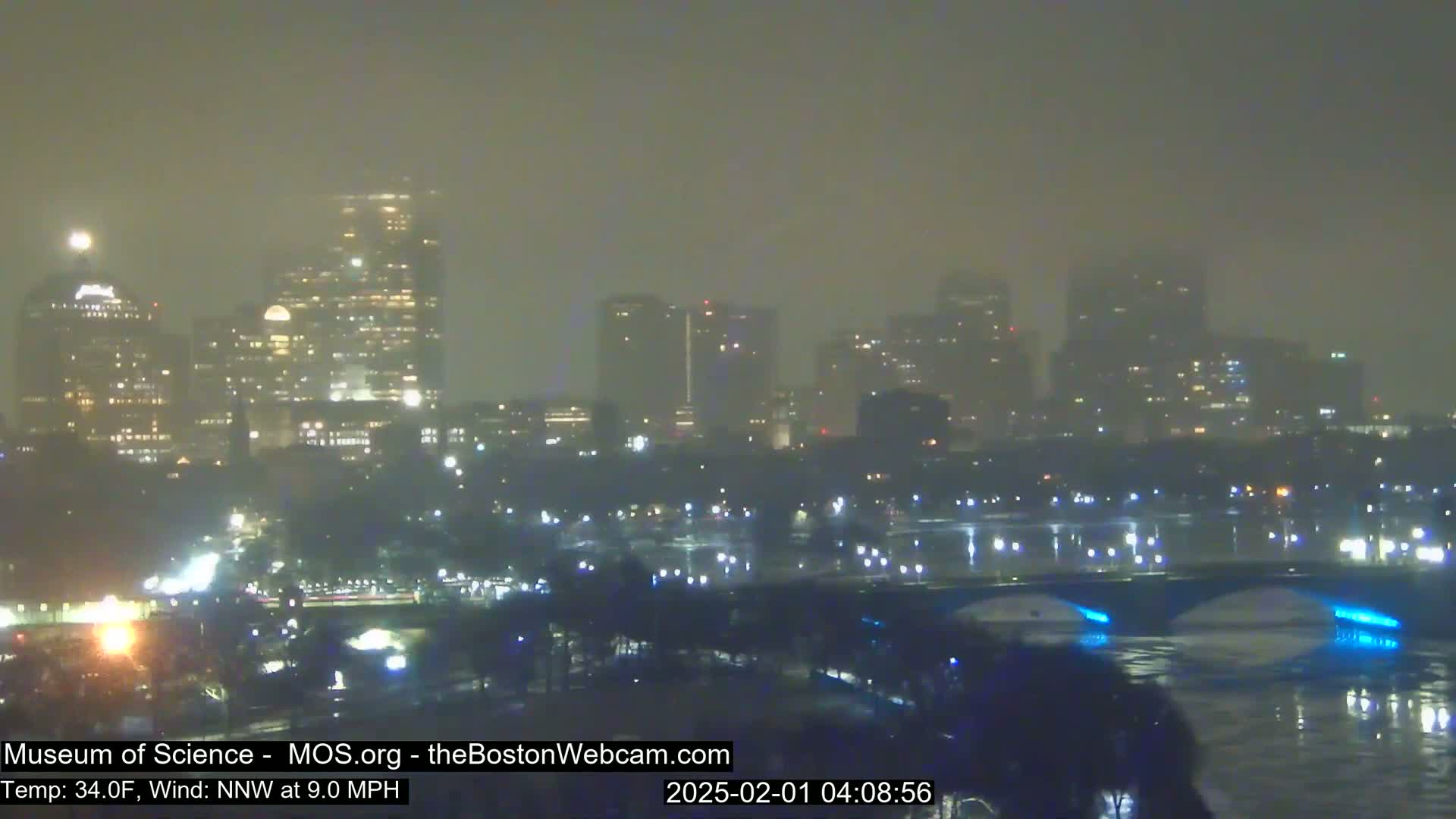 A hazy, nighttime view of a city skyline across a body of water with a bridge illuminated in blue.