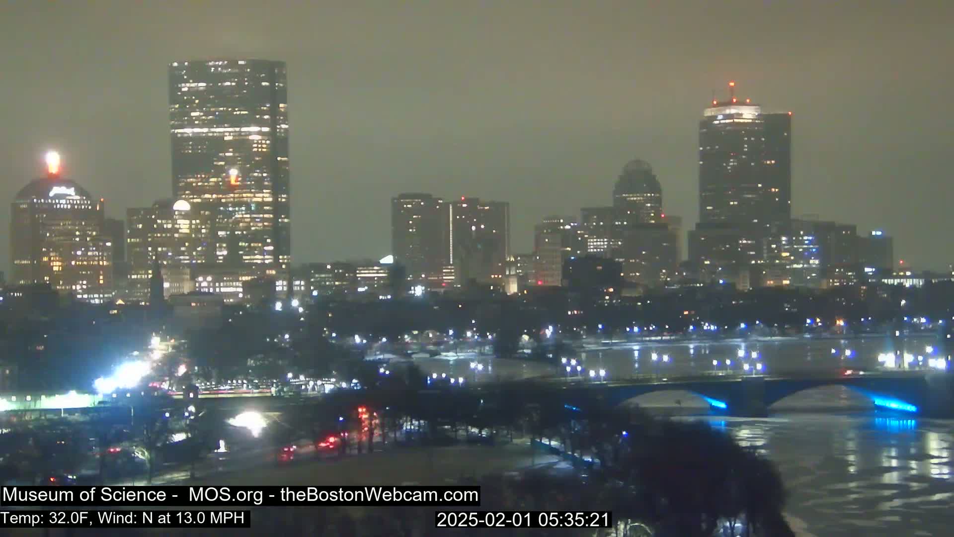A nighttime cityscape with several illuminated skyscrapers and a bridge over a partially ice-covered river under hazy conditions.