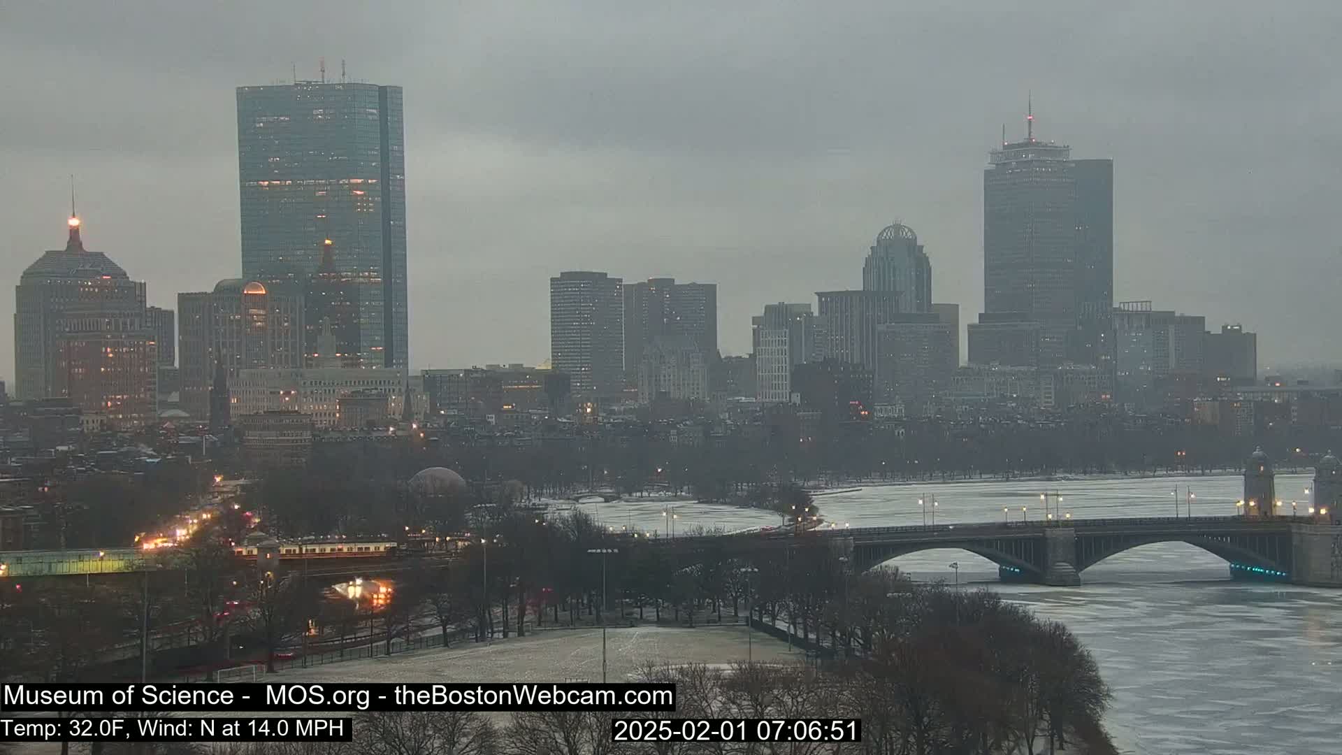 A city skyline with several tall buildings is visible across a partially frozen river under an overcast sky.