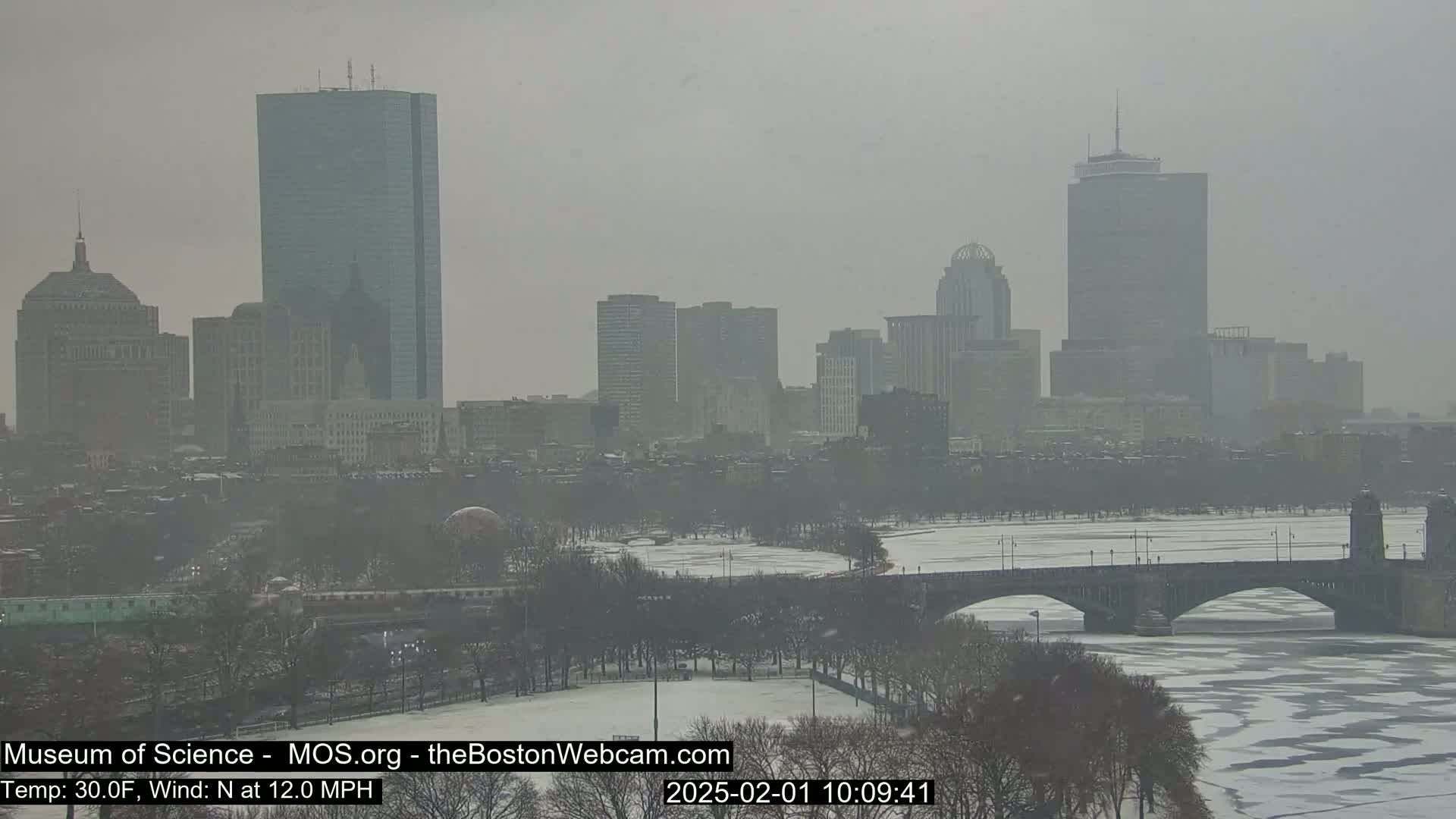 A hazy, snowy cityscape with a partially frozen river and a bridge in the foreground.
