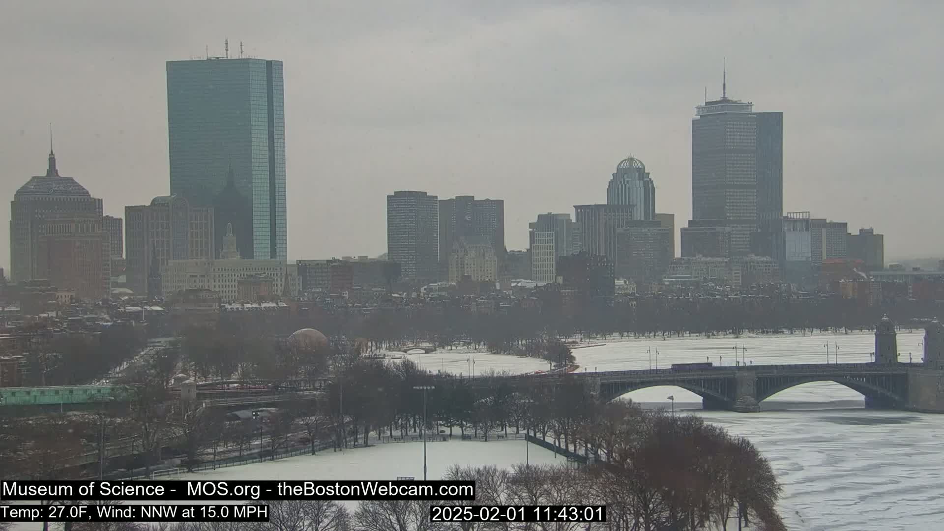 A snow-covered cityscape, including several tall buildings and a partially frozen river with a bridge crossing it, is visible under an overcast sky.