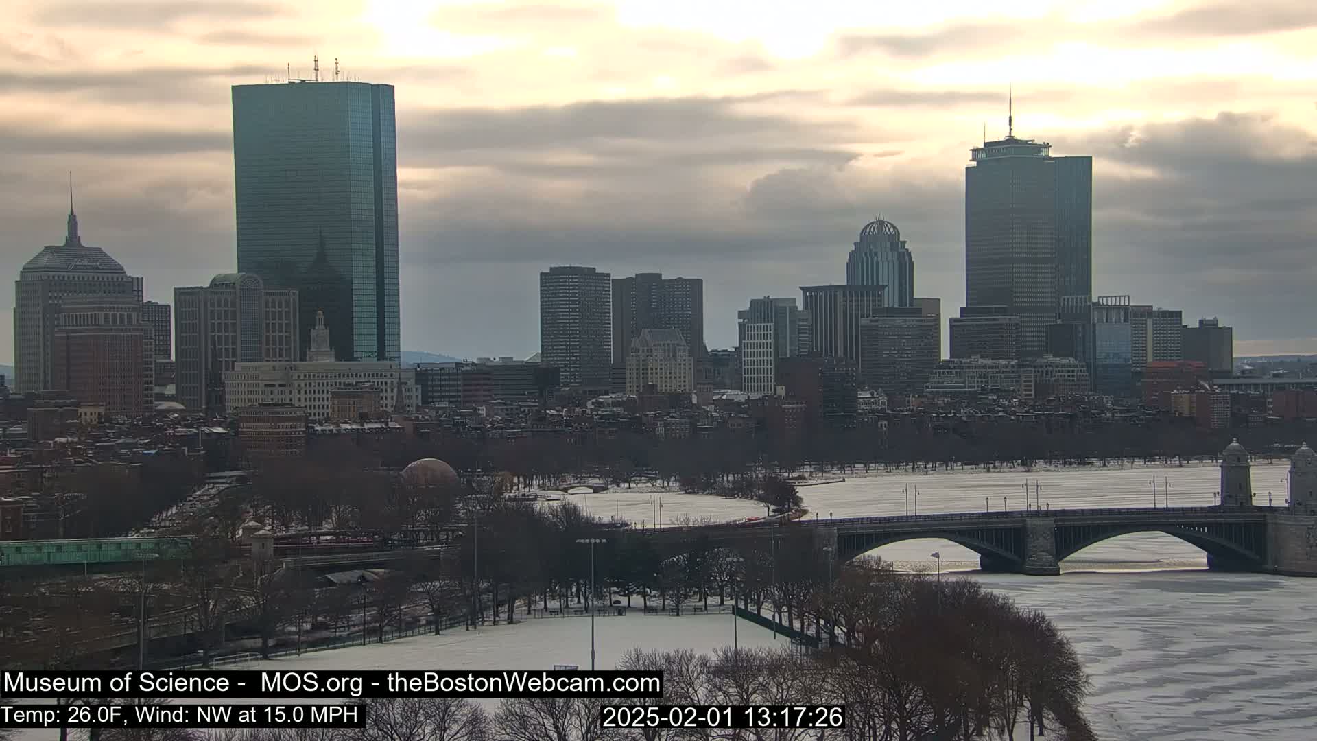 A snow-covered Boston cityscape with a partly frozen river and bridge under a cloudy sky.