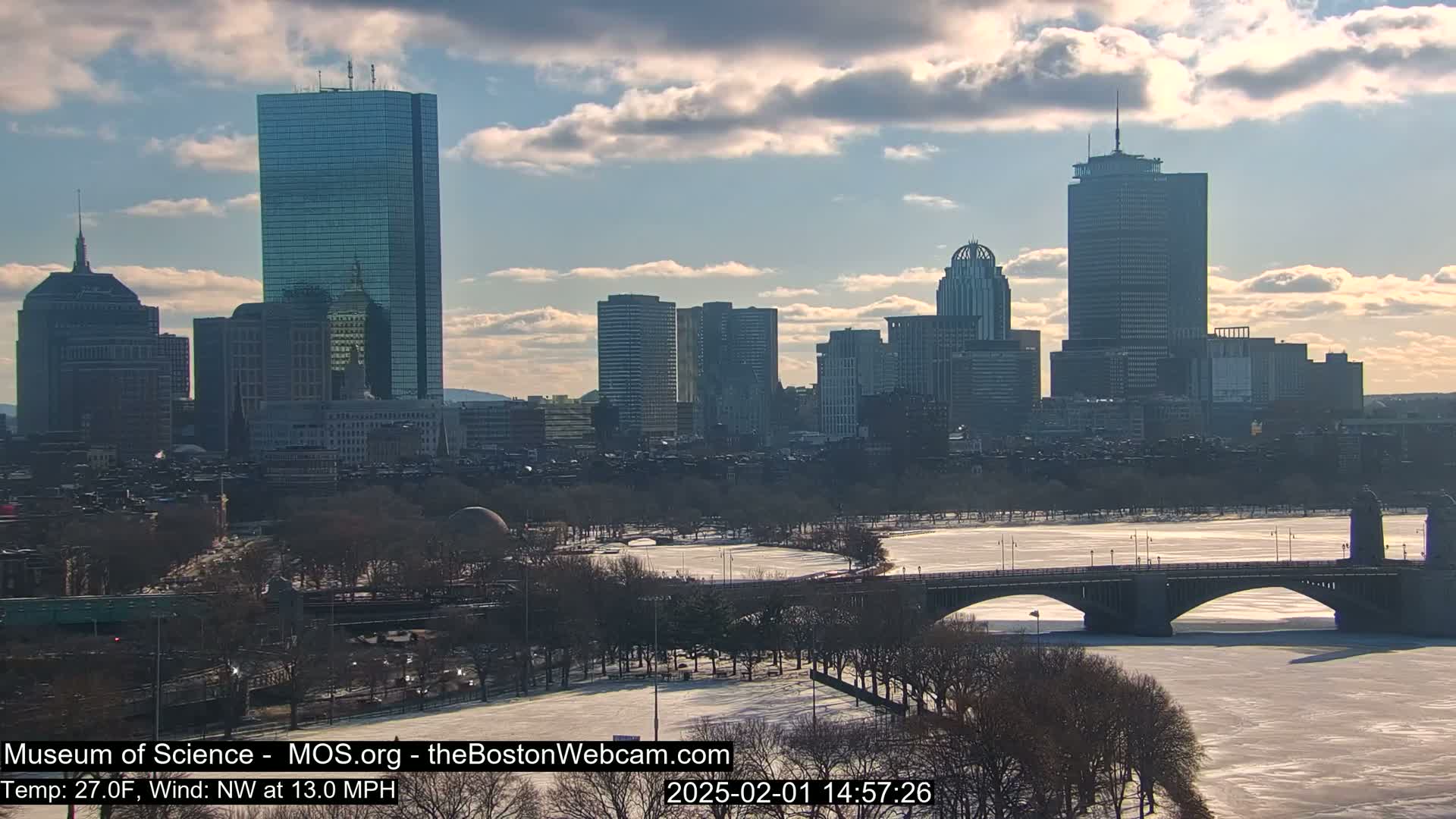 A partly cloudy winter day shows a city skyline across a partially frozen river with a bridge; the trees are bare.