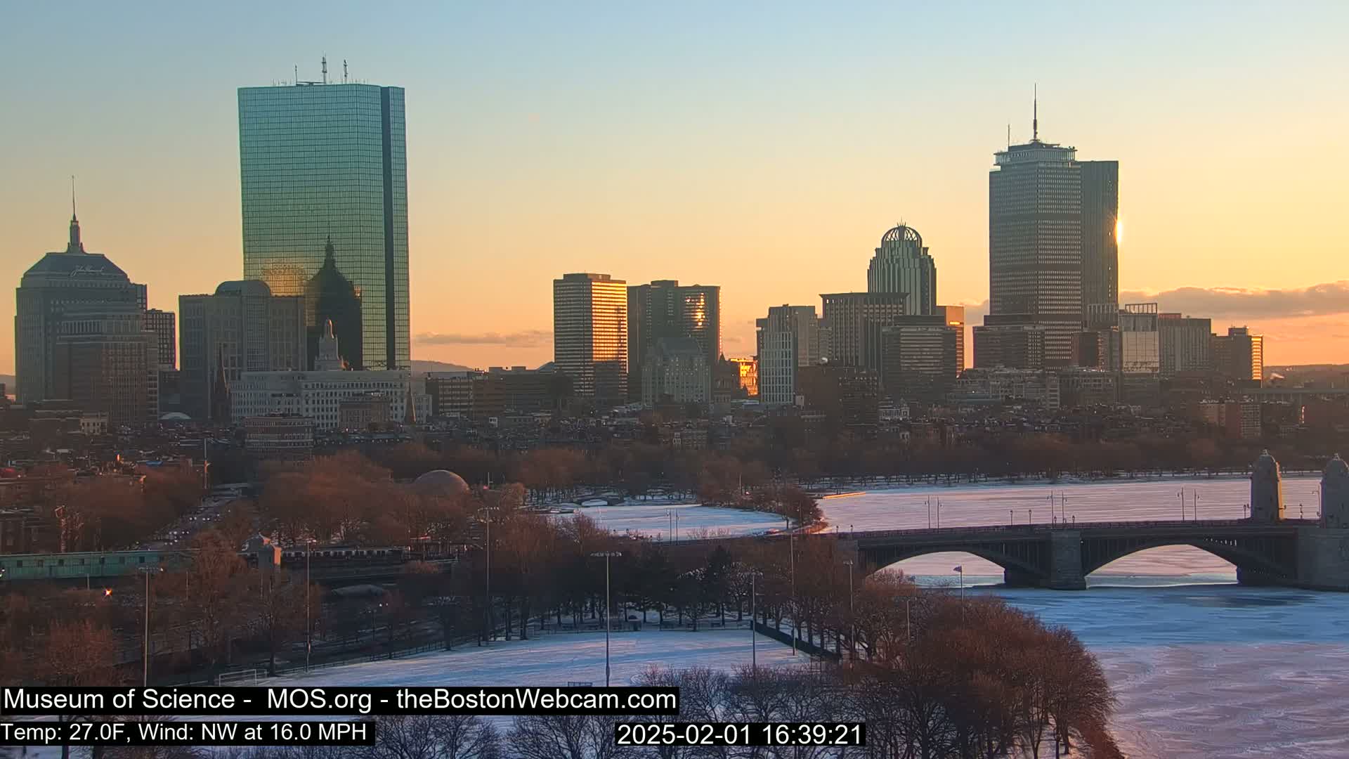 A city skyline at sunset, with a partially frozen river and bridge in the foreground, under a clear, twilight sky.