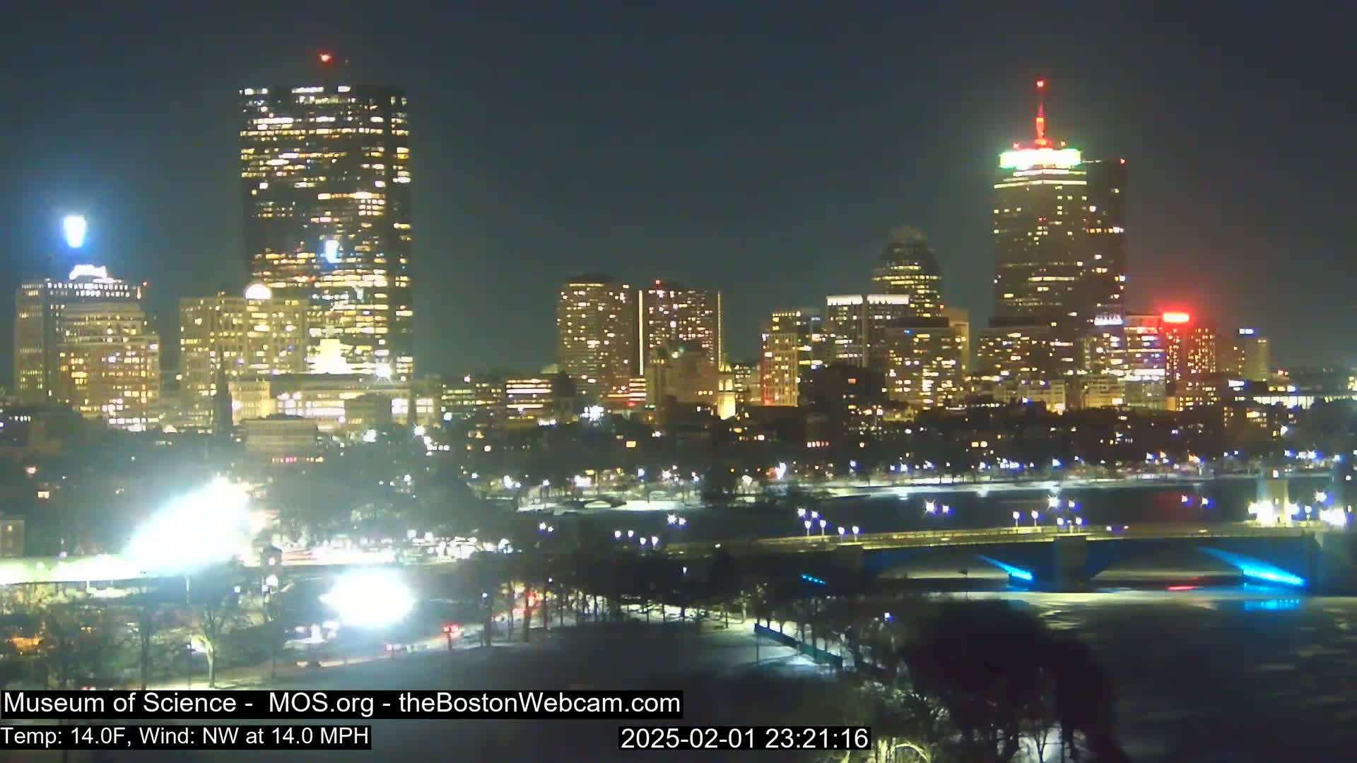 A nighttime cityscape with numerous illuminated buildings, a bridge, and a body of water under a clear sky, with a temperature of 14°F and a northwest wind of 14 mph.