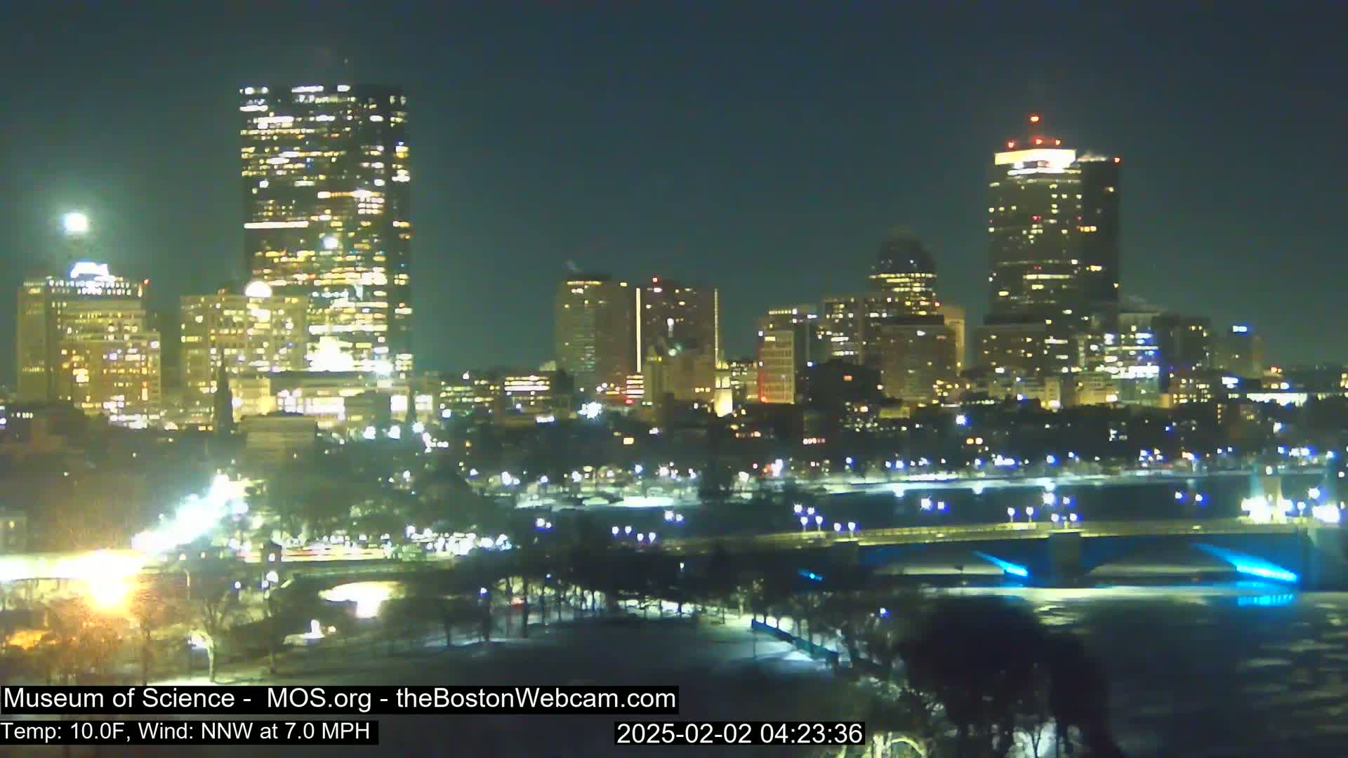 A nighttime view of a city skyline illuminated by lights, with a river in the foreground and snow on the ground, under a clear sky.