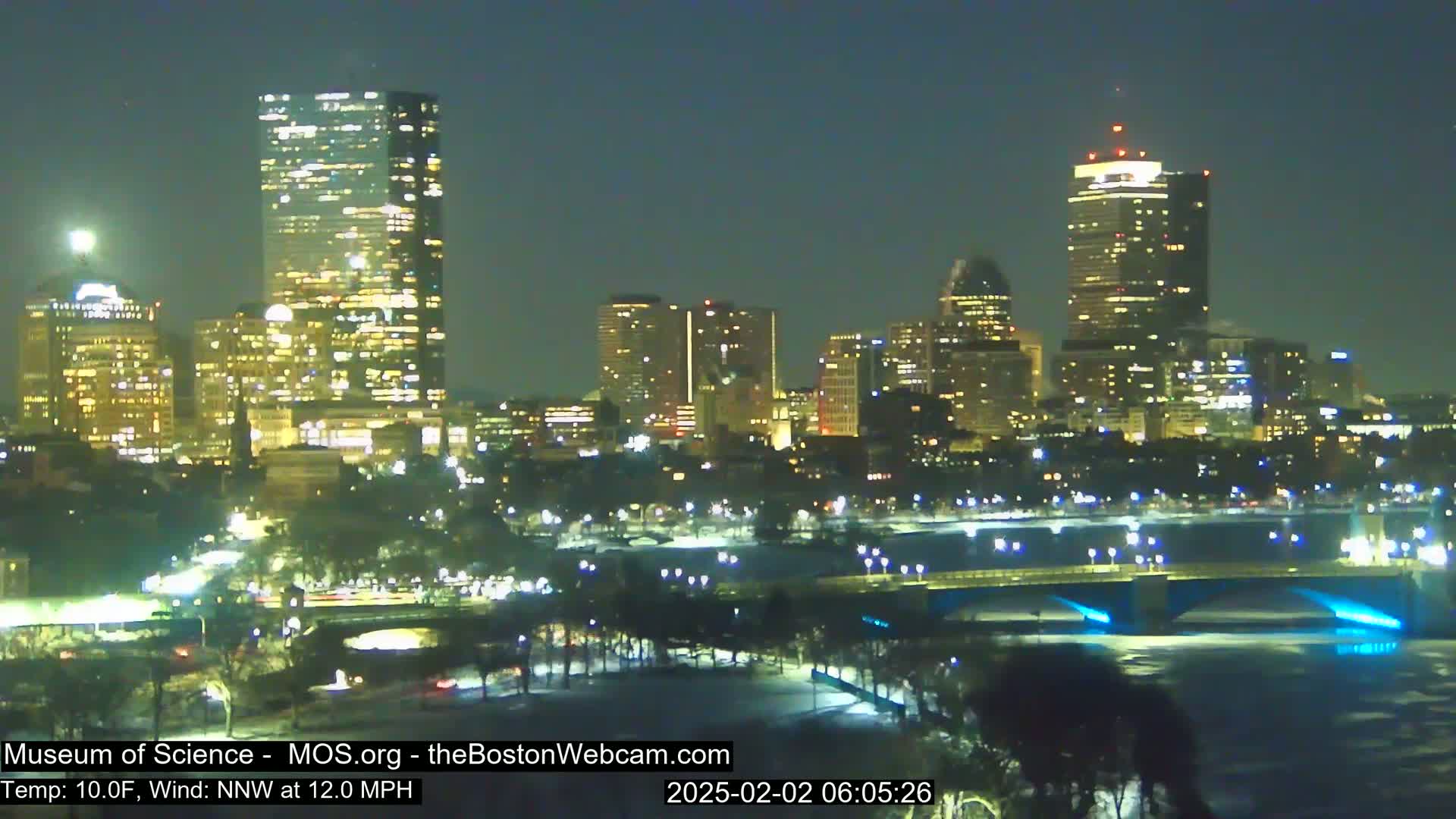 A nighttime view of a city skyline illuminated by lights, with a bridge spanning a partially frozen river under a clear, moonlit sky and a northwest wind.