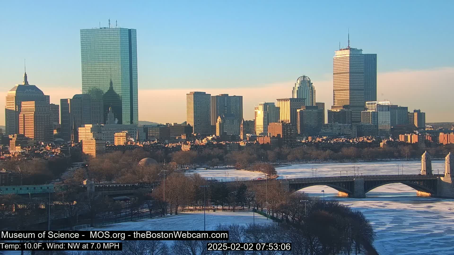 A cityscape at sunrise shows a river partially frozen over, a bridge, and numerous buildings under a clear sky.