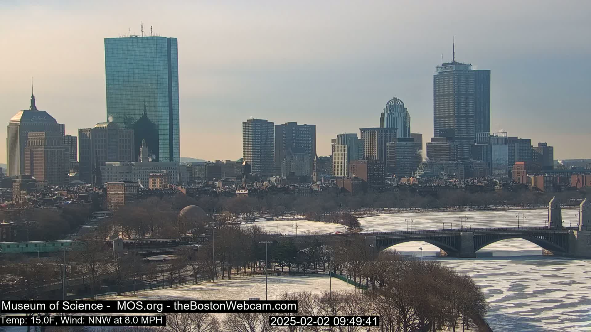 A partially frozen river flows beneath a bridge in front of a cityscape on a partly sunny, cold day.