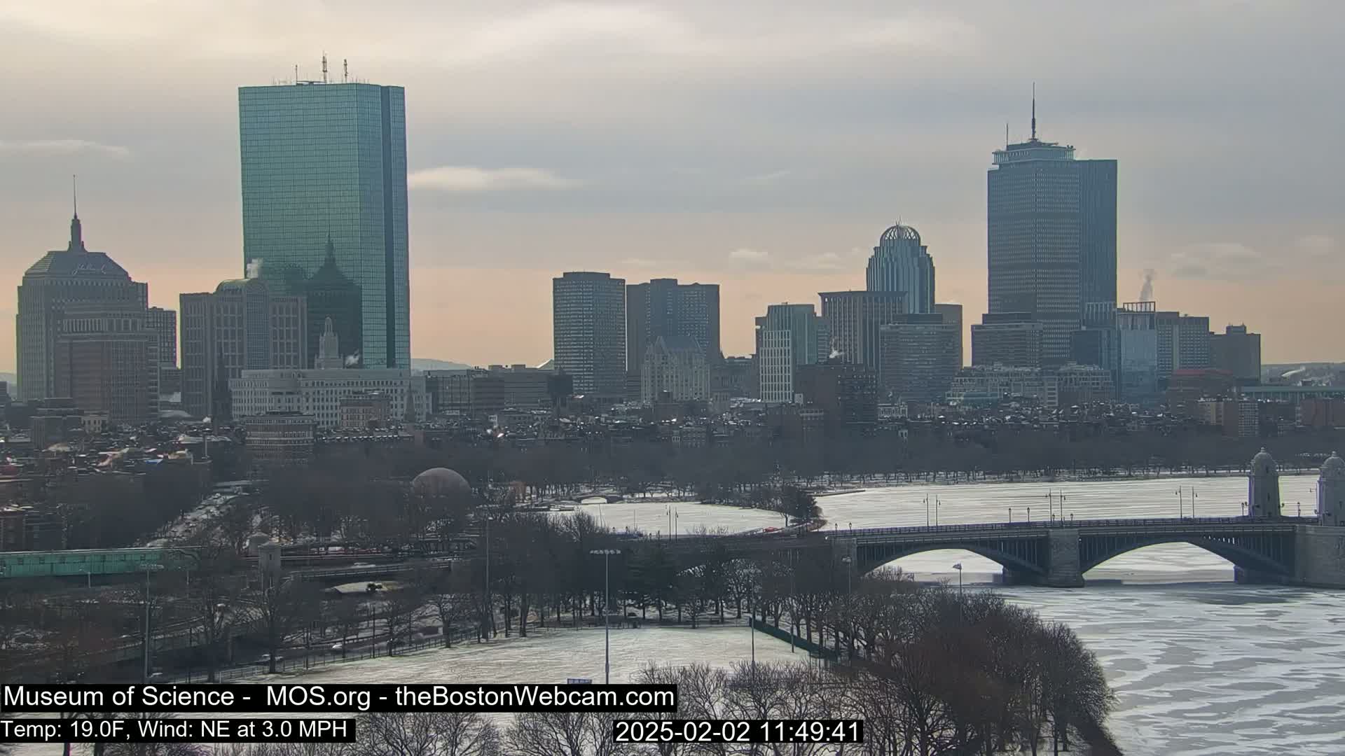 A snow-covered cityscape with a partially frozen river and a bridge under a cloudy sky.
