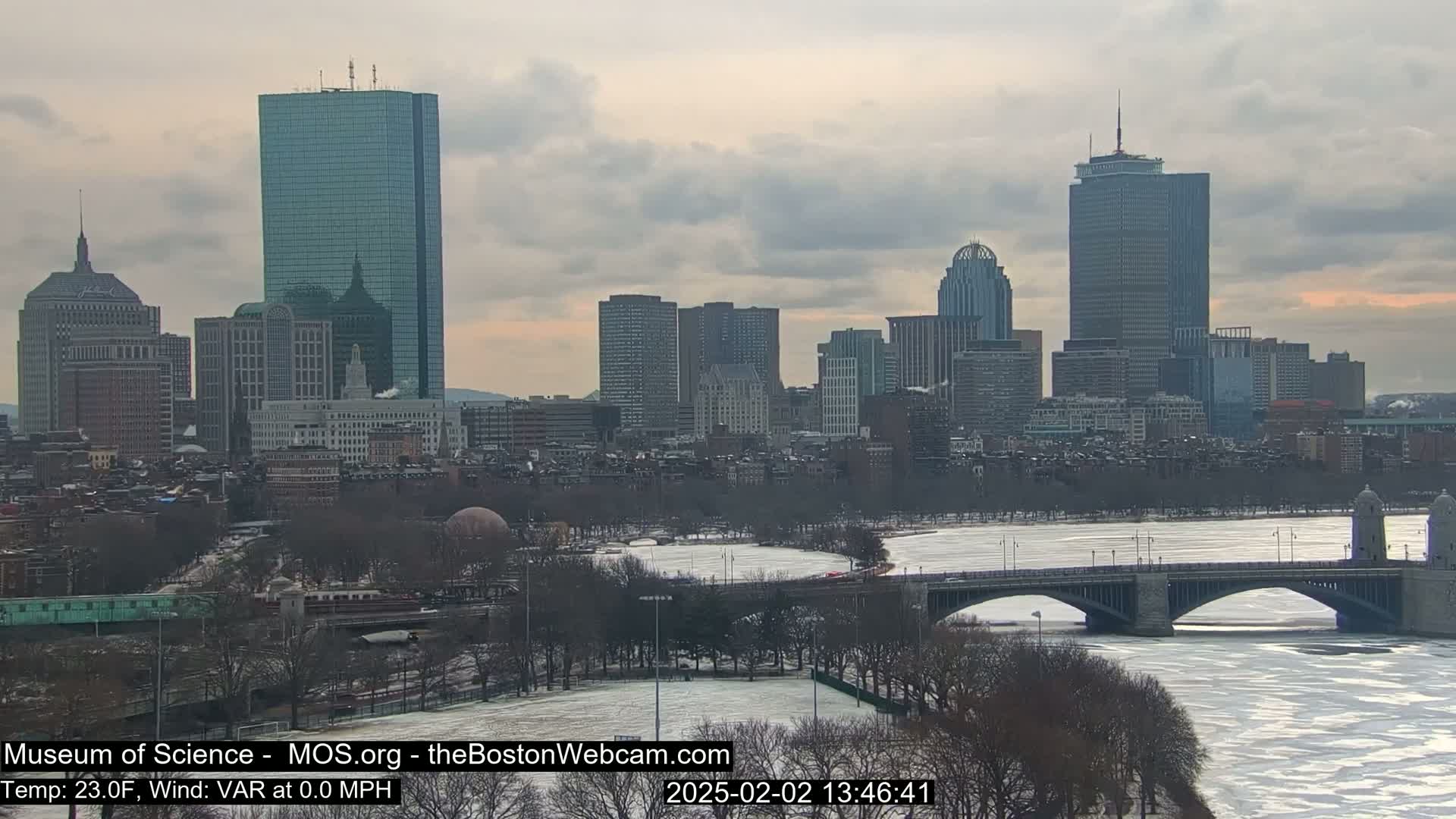 A partially frozen river flows beneath a bridge in front of a city skyline under a cloudy sky.
