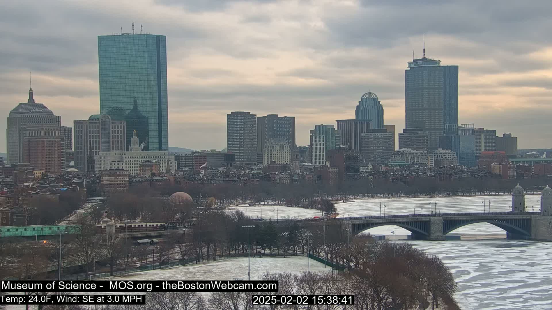 A partially frozen river flows beneath a bridge in front of a cityscape under a cloudy sky.