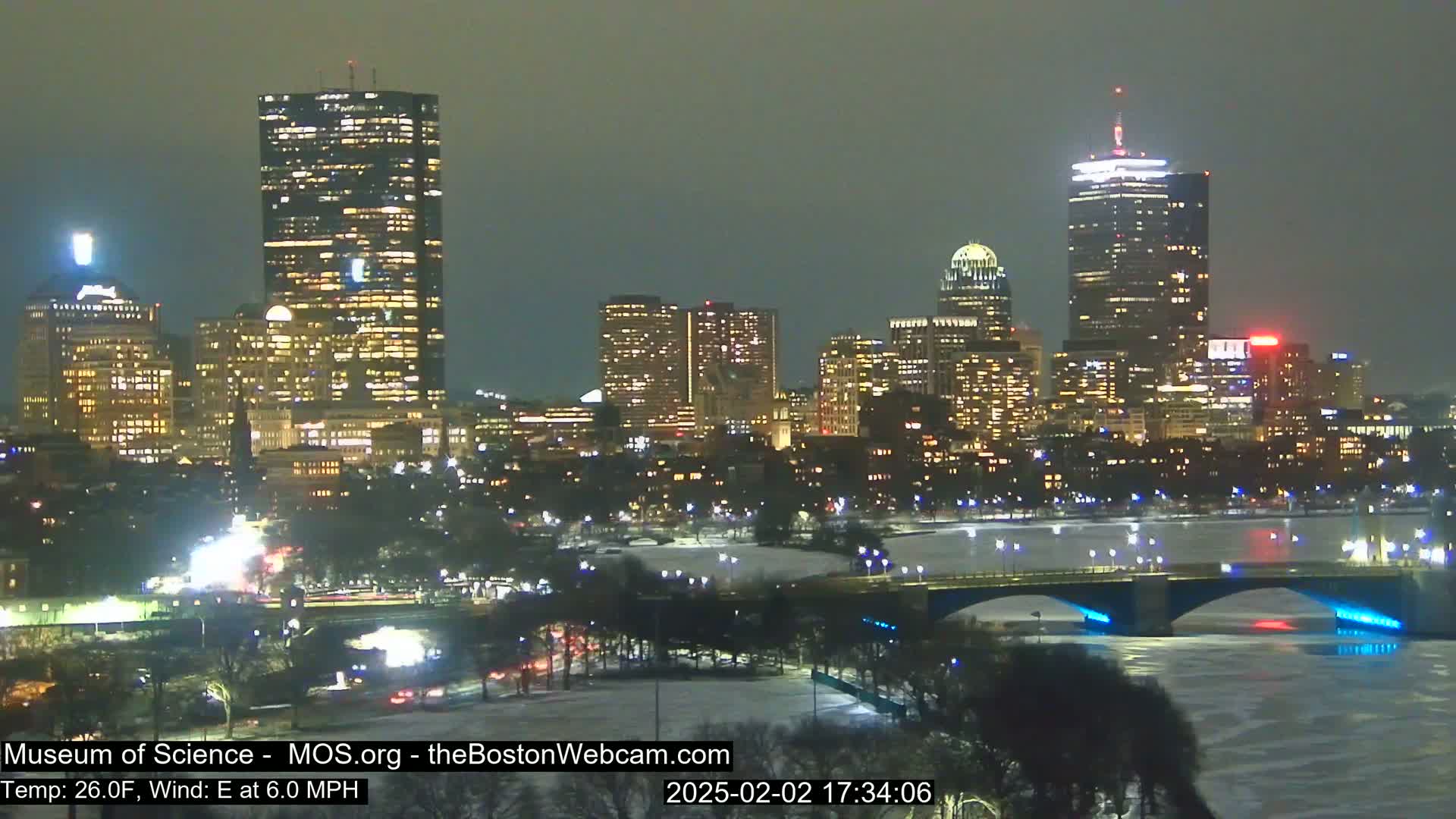A nighttime view of a city skyline illuminated with lights across a partially frozen river and bridge, under a dark sky.