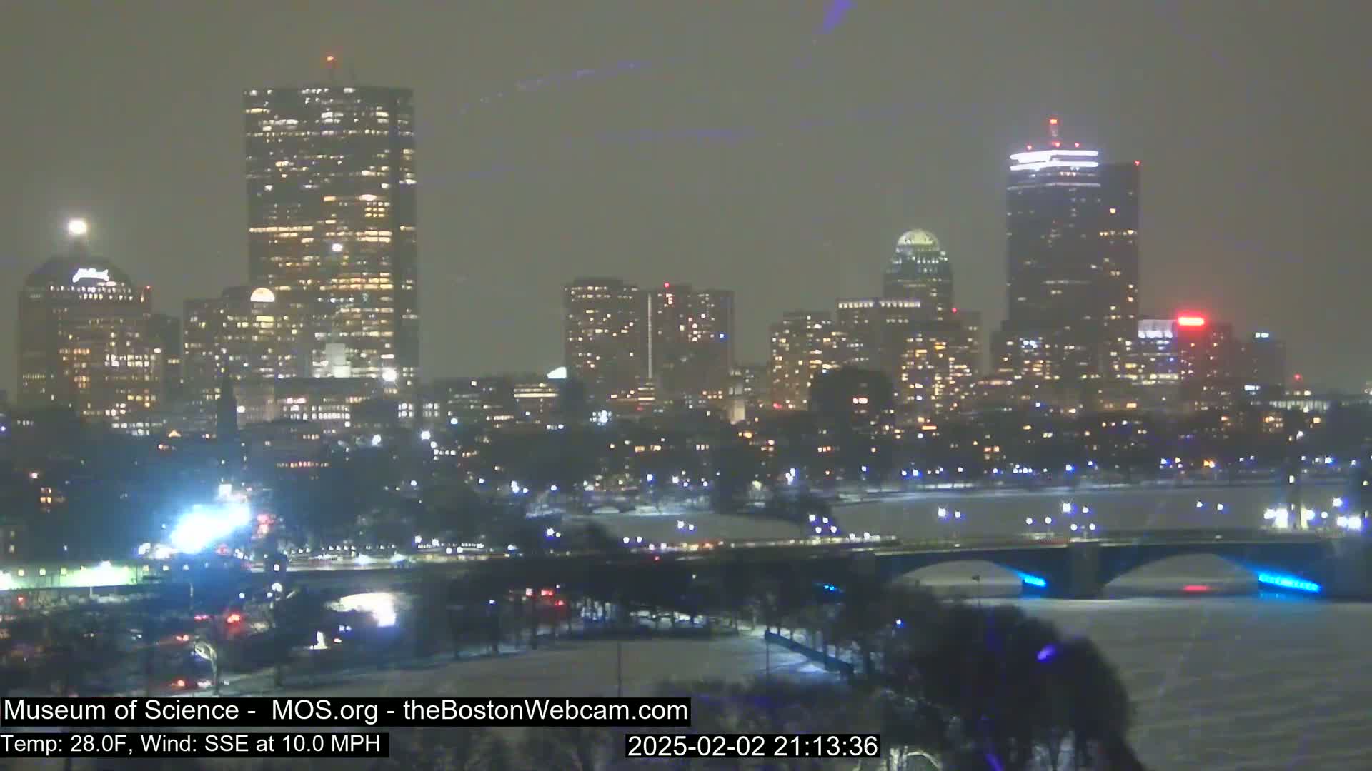 A nighttime view of a snow-covered city skyline with a bridge and river, under a slightly overcast sky.