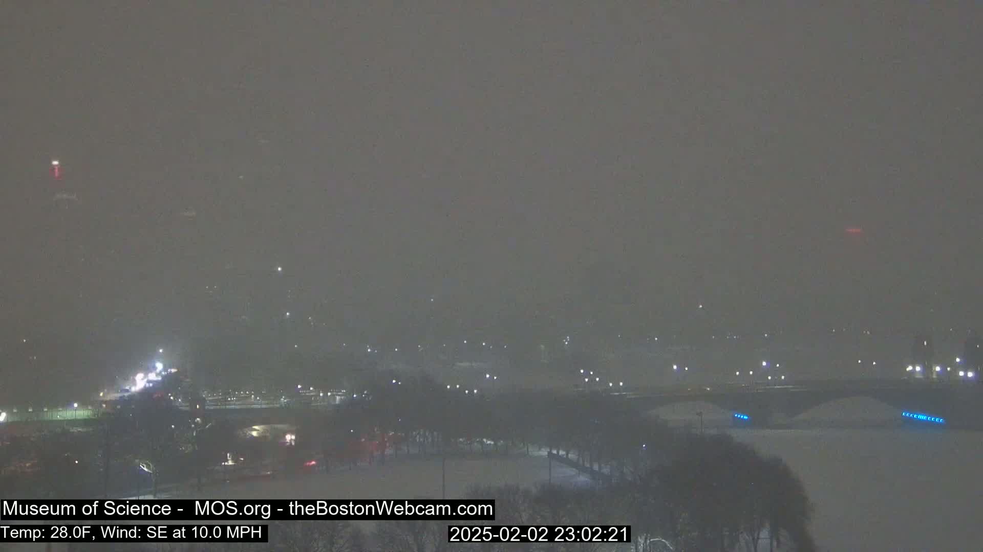 A snow-covered landscape at night, with faint lights visible in the distance, under heavy snowfall and a southeast wind.