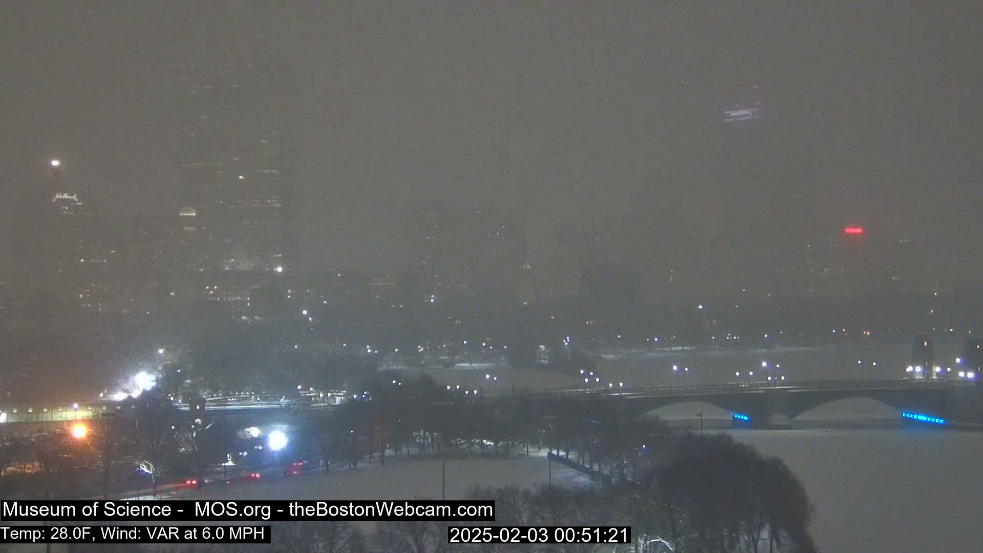 A snow-covered cityscape at night, with a bridge spanning a snow-covered river, is visible through heavy snowfall.