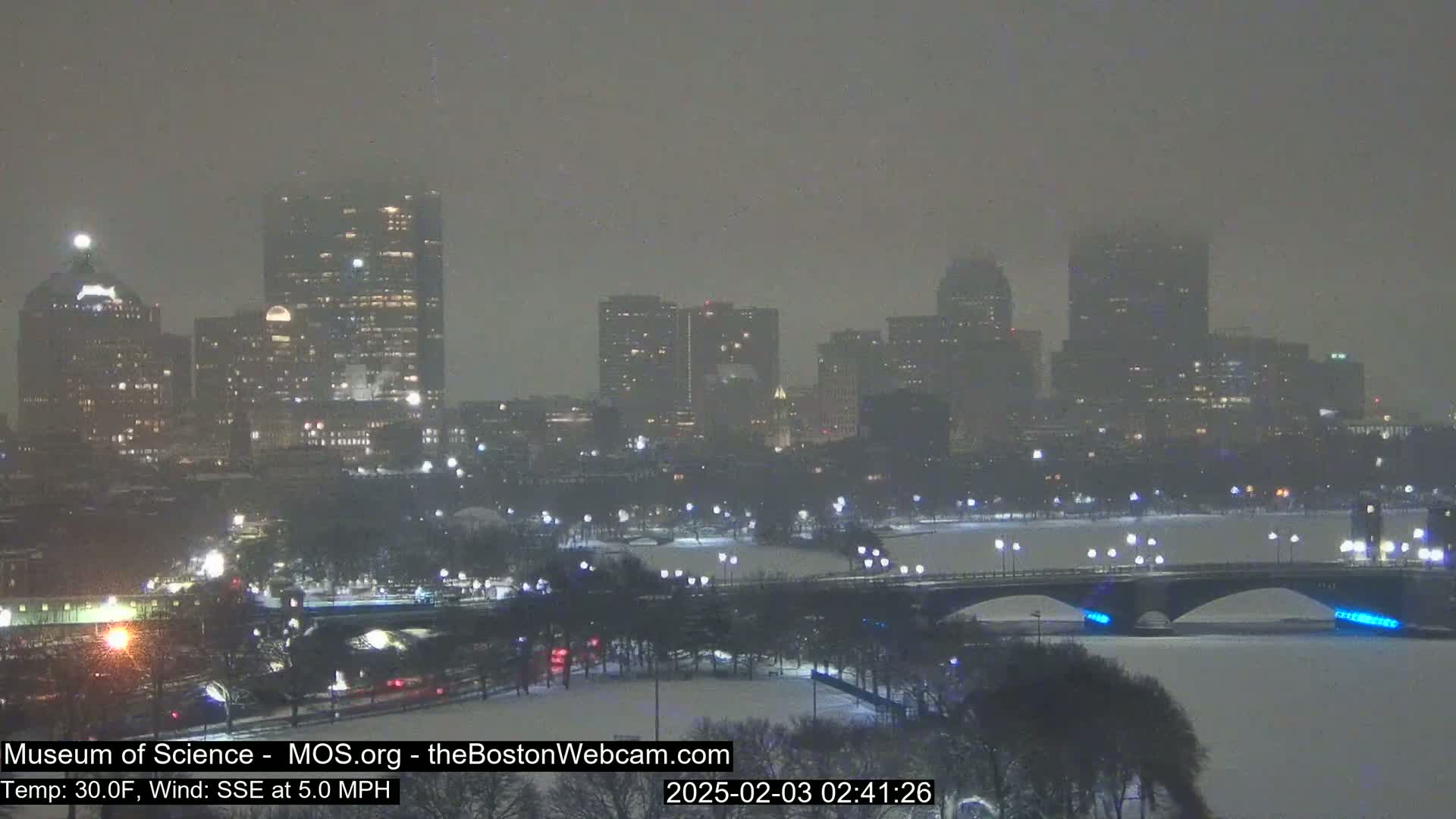 A snow-covered city skyline with illuminated buildings is viewed across a river and bridge at night during a snowstorm.