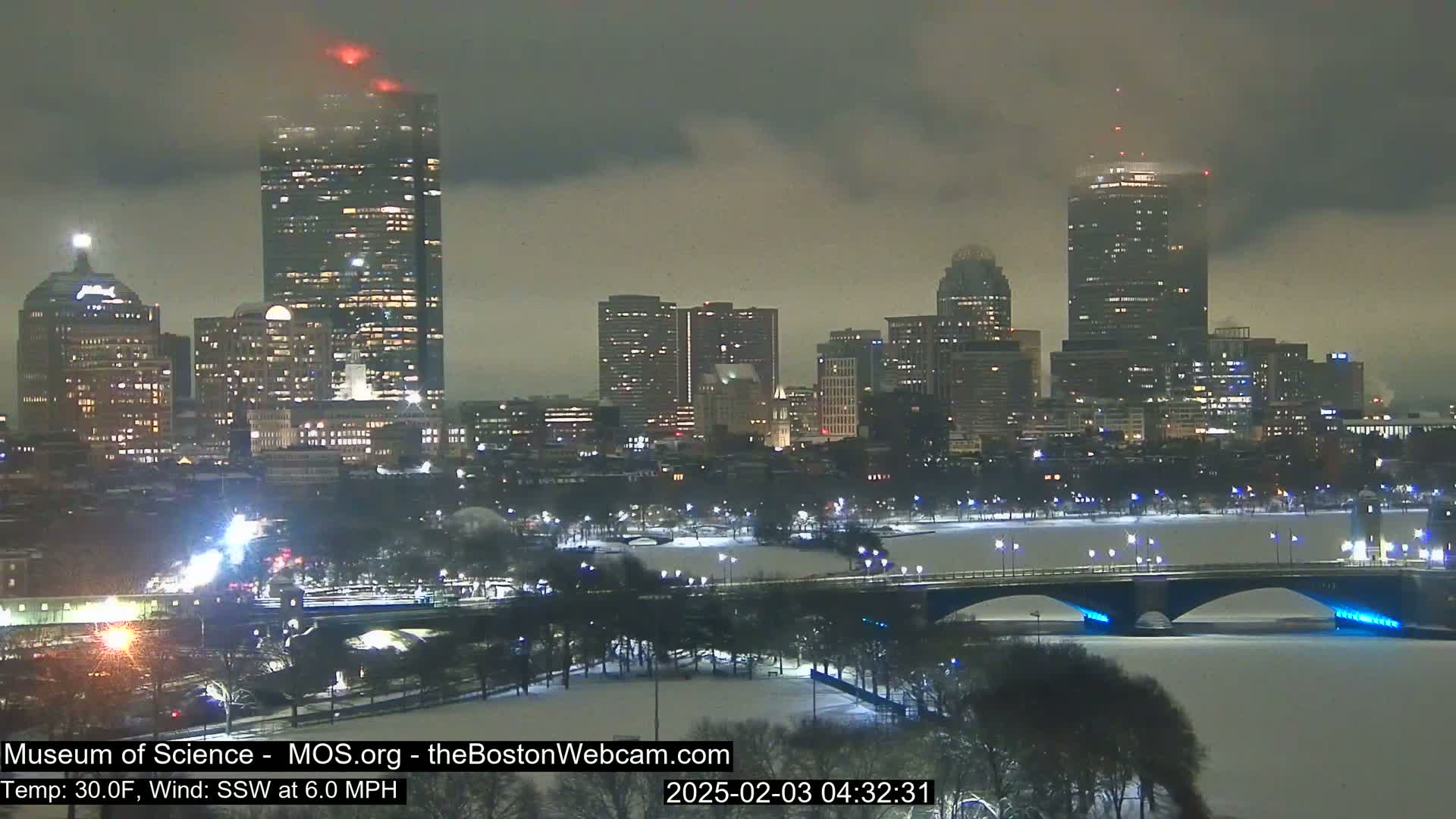 A snow-covered cityscape at night with a bridge and river, under a cloudy, dark sky.