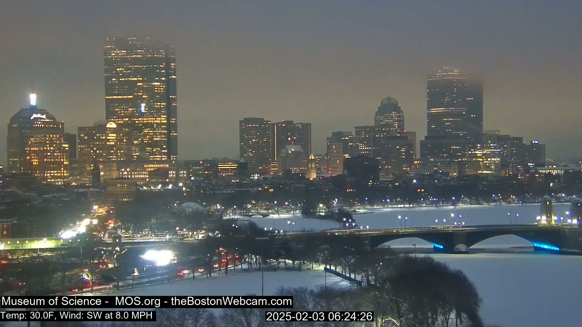 A snow-covered city skyline at night, illuminated by building lights, with a bridge spanning a frozen river in the foreground under a dusky sky.