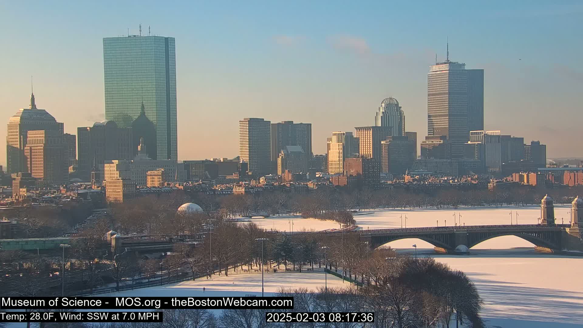 A snow-covered cityscape with a frozen river and bridge under a clear, sunny sky.