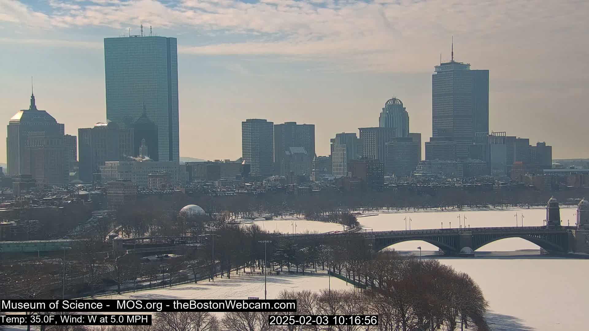 A snow-covered cityscape with a bridge over a partially frozen river under a partly cloudy sky.