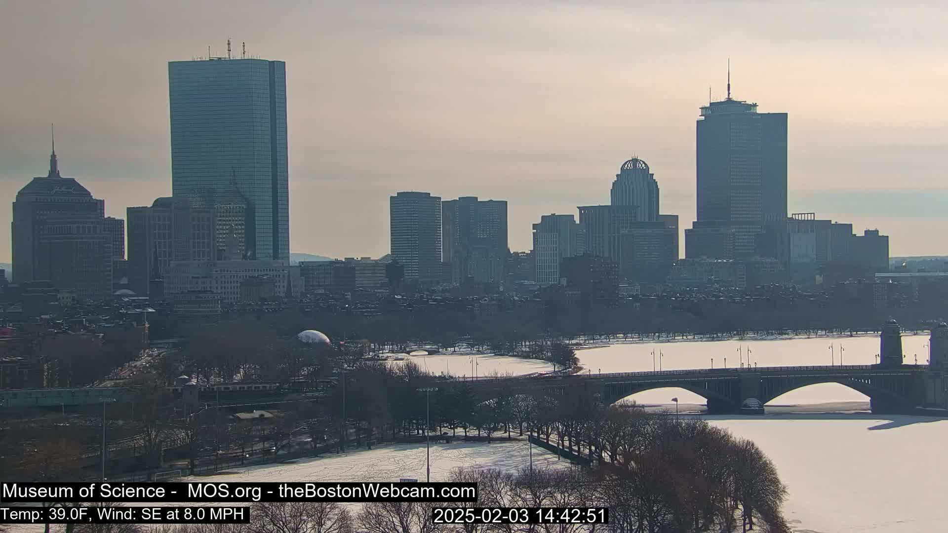 A snow-covered city skyline with several skyscrapers, a bridge over a partially frozen river, and leafless trees under a mostly clear sky.