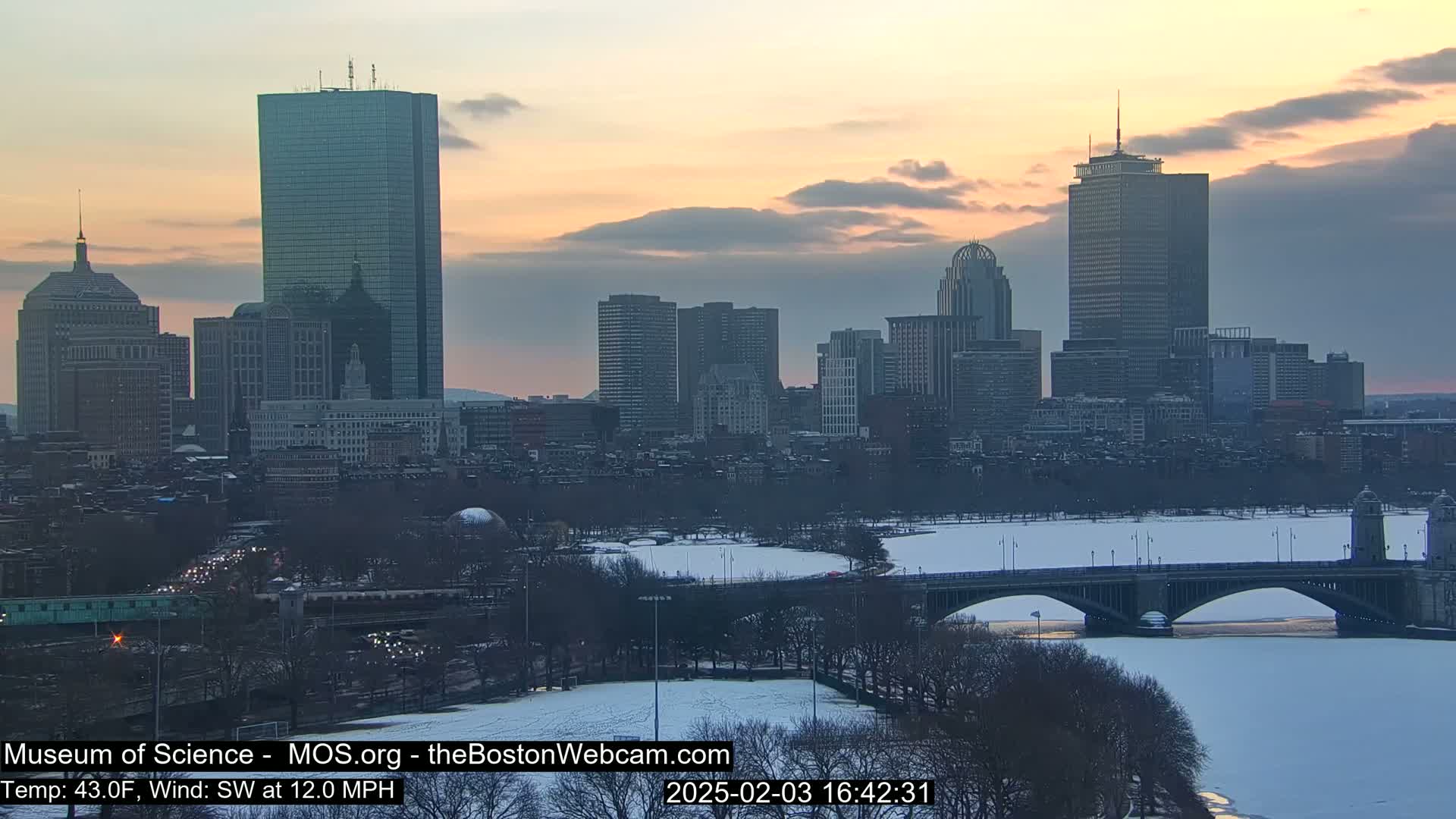 A city skyline at sunset with a partly frozen river and bridge underneath, under partly cloudy conditions.