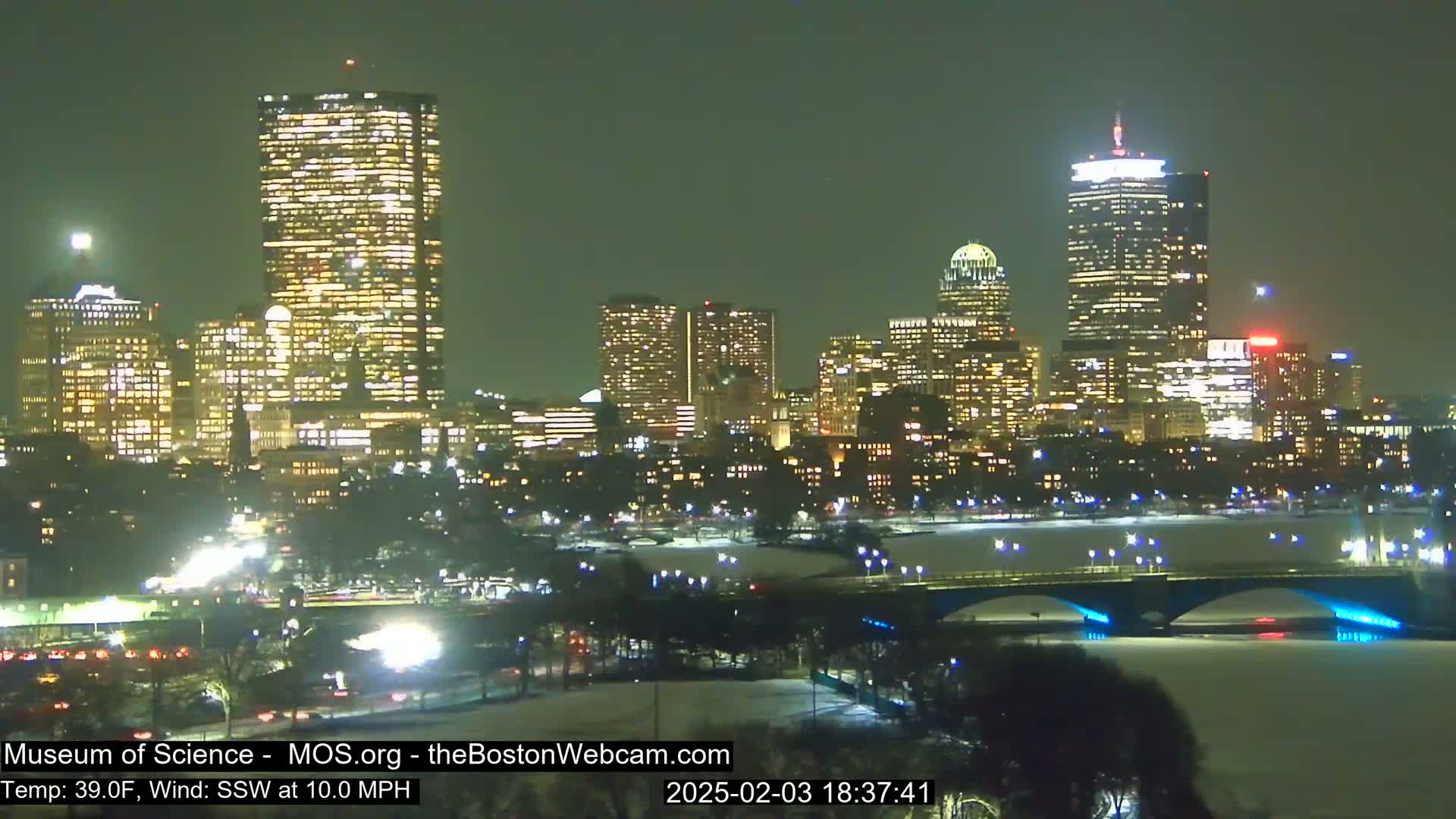 A nighttime cityscape with illuminated skyscrapers and a bridge over a snow-covered river, under a partly cloudy sky with a 10 mph southwest wind and a temperature of 39°F.