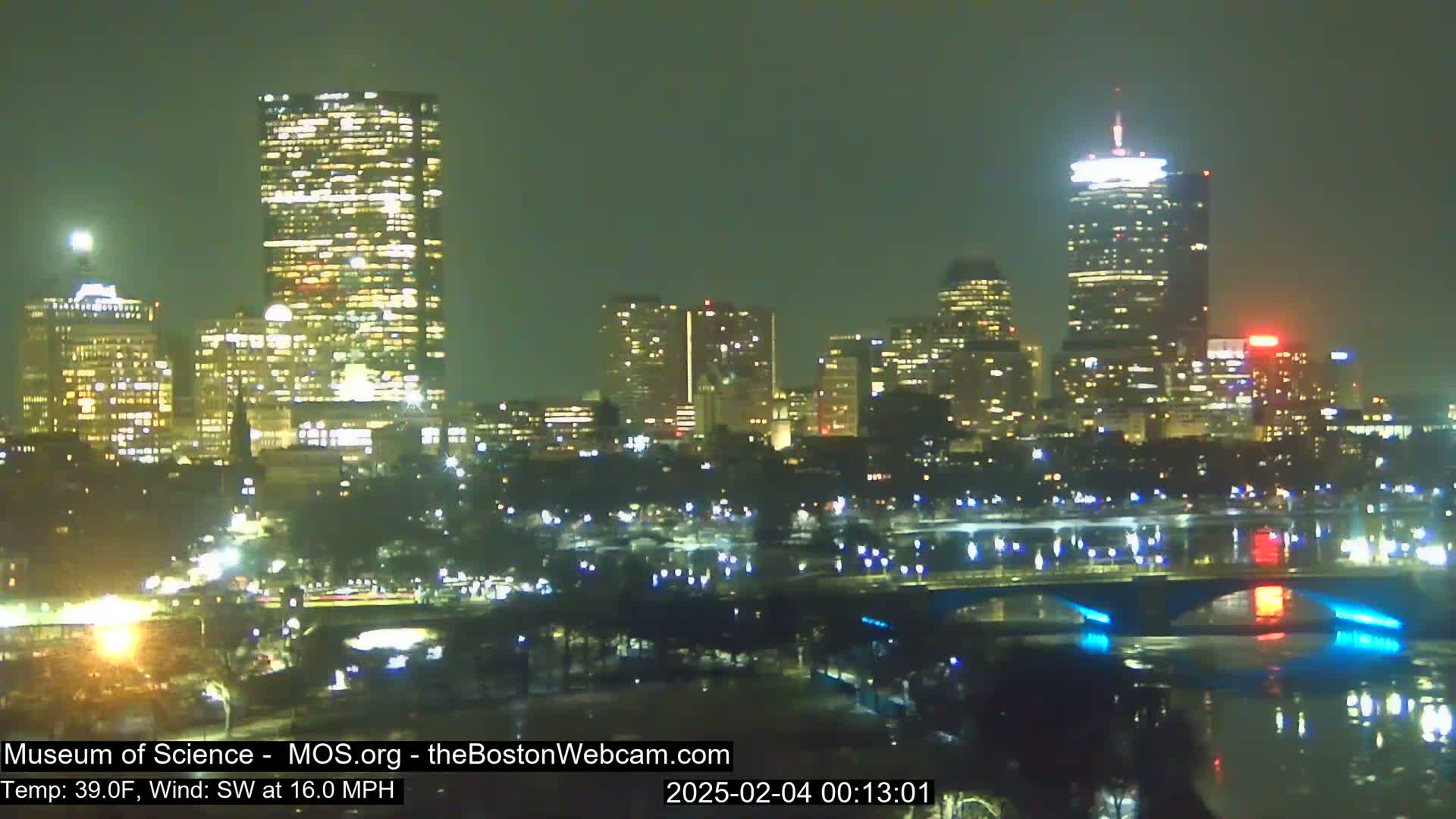 A nighttime view of a city skyline across a body of water, under a clear, dark sky with a southwest wind at 16 mph and a temperature of 39°F.