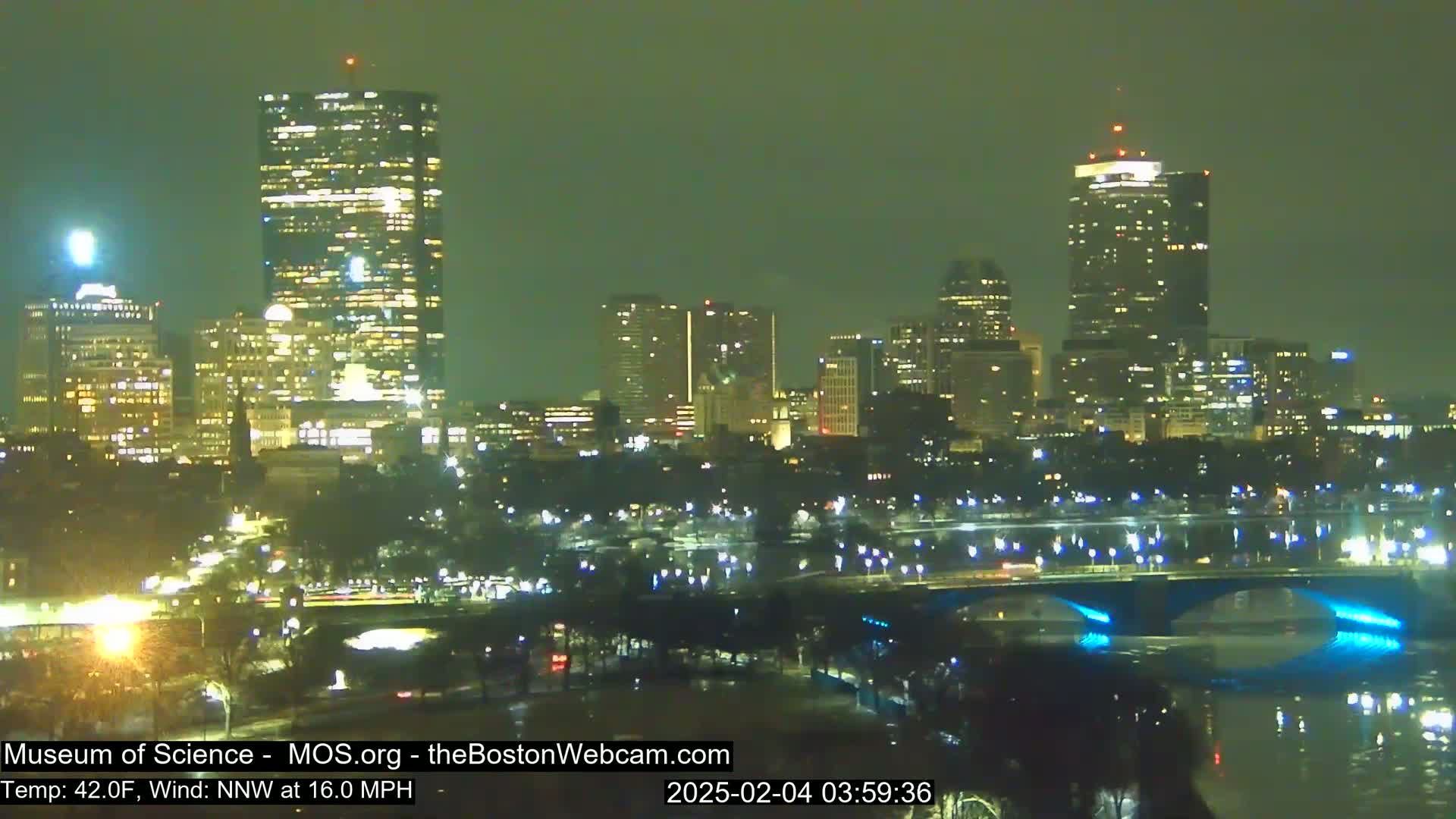 A nighttime view of a city skyline illuminated by lights, with a bridge spanning a body of water under a dark, possibly cloudy sky, and a wind of 16 mph.