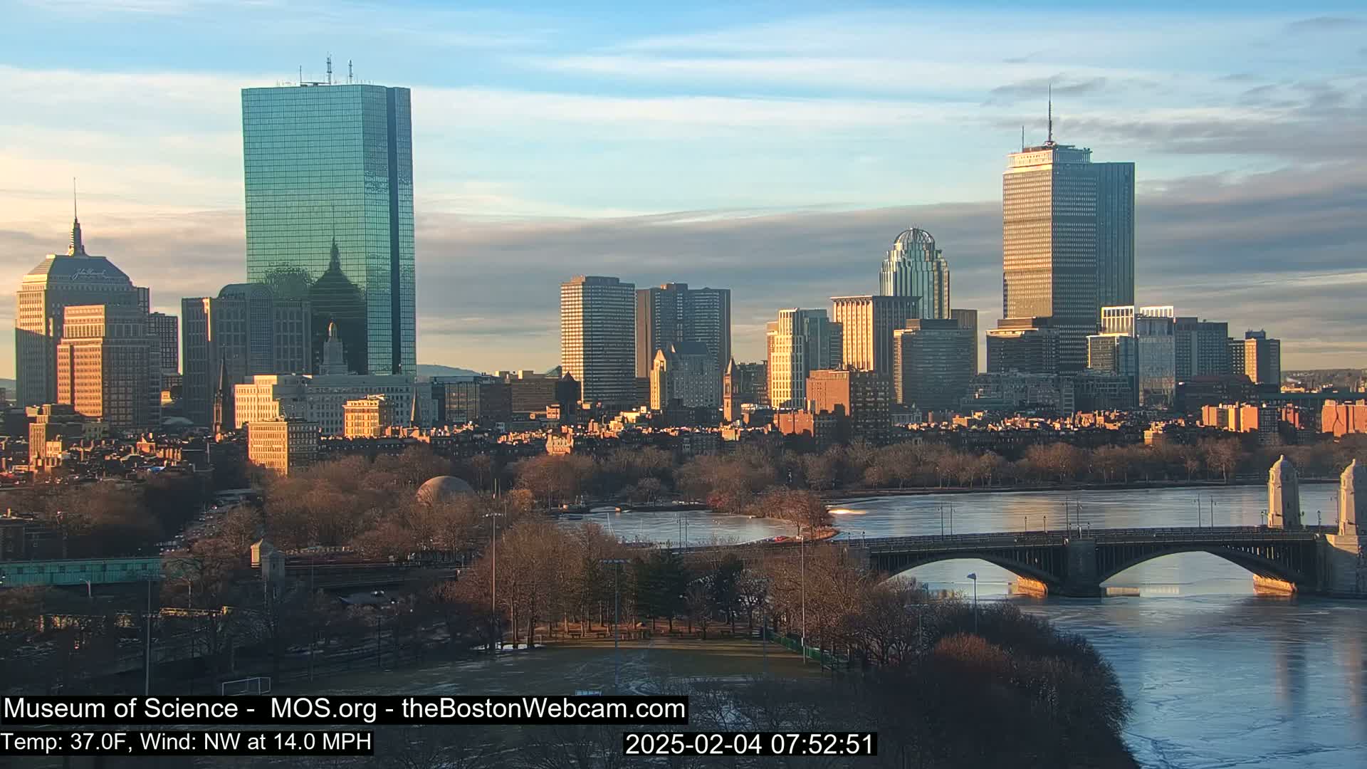 A city skyline with numerous skyscrapers is visible across a partially frozen river, under a partly cloudy sky at sunrise.