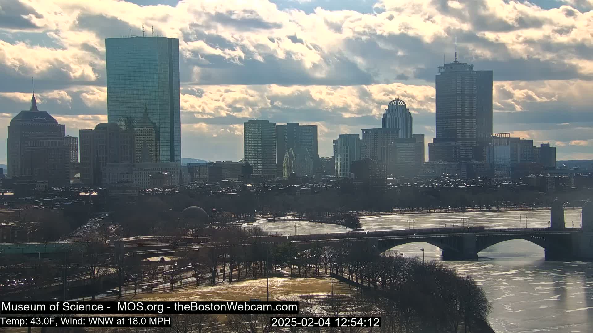 A city skyline with several tall buildings is visible under a partly cloudy sky, with a partially frozen river and a bridge in the foreground.
