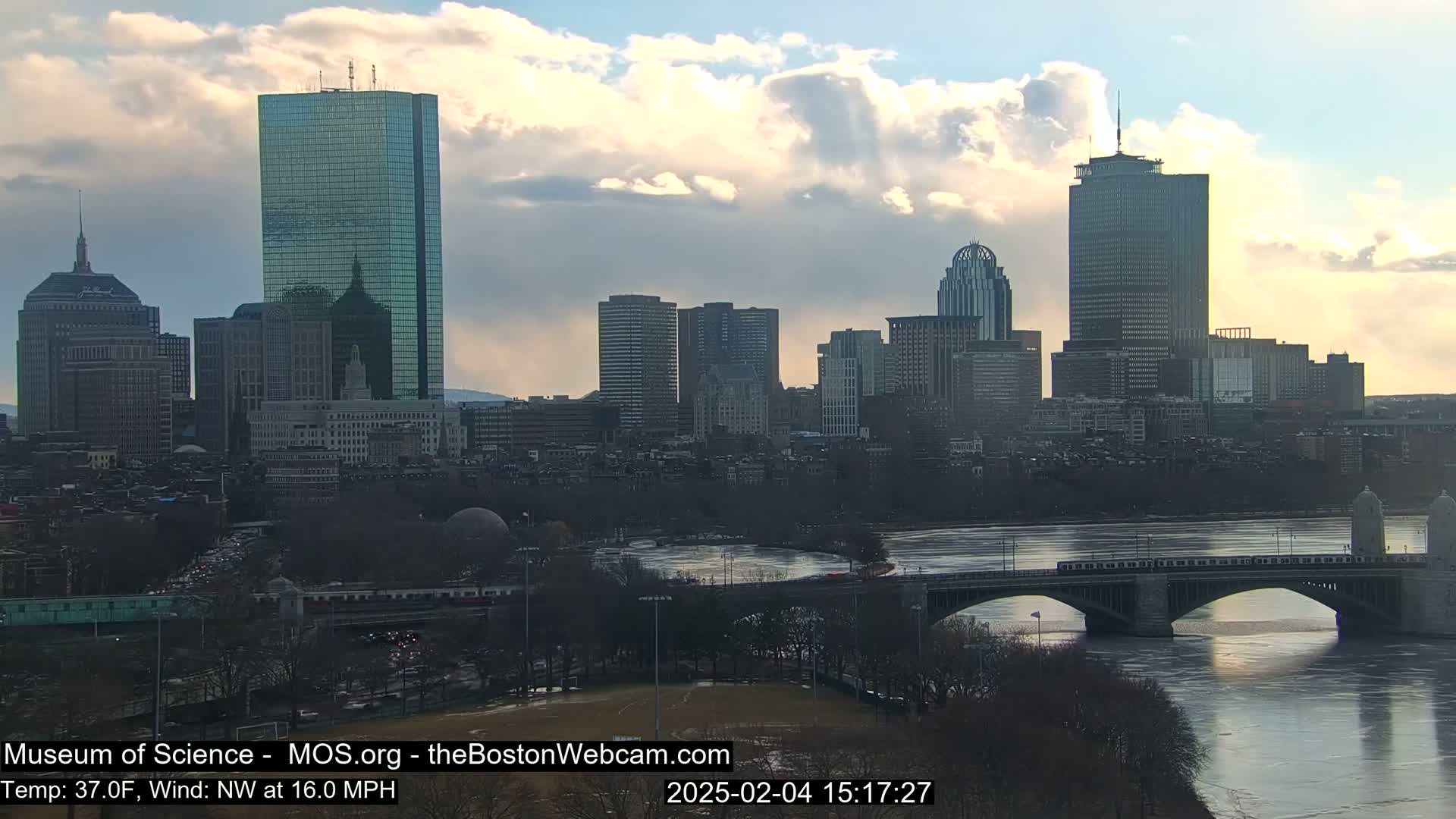 A city skyline with numerous skyscrapers is visible across a river with a bridge, under a partly cloudy sky.