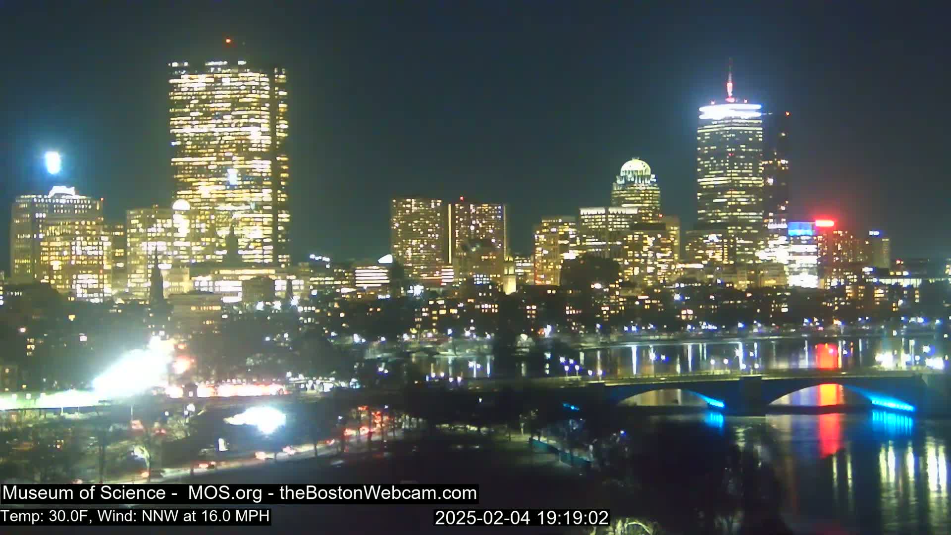 A brightly lit city skyline at night, viewed from across a river with a bridge, under a clear night sky with a 30°F temperature and a 16 mph north-northwest wind.