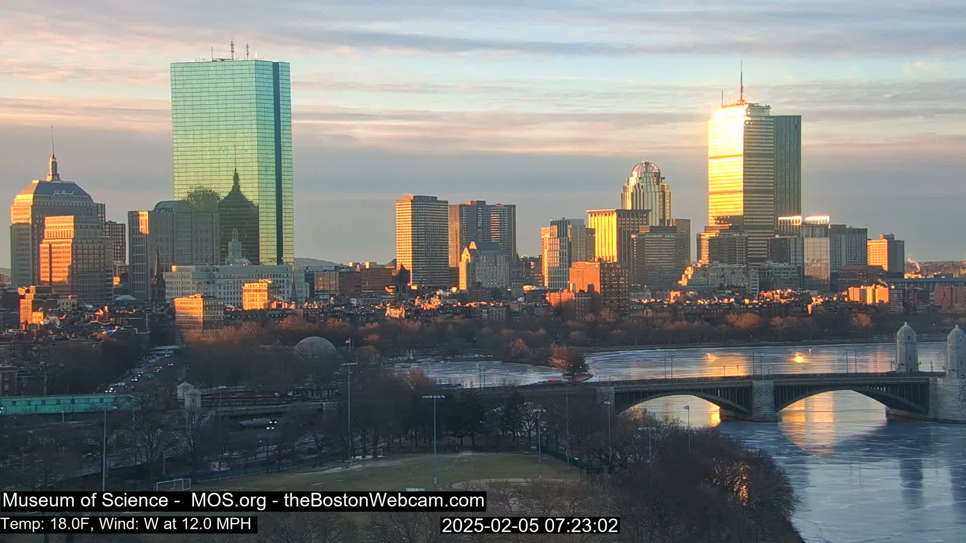 A city skyline at sunrise, with a partly frozen river and bridge in the foreground, under a mostly clear sky.