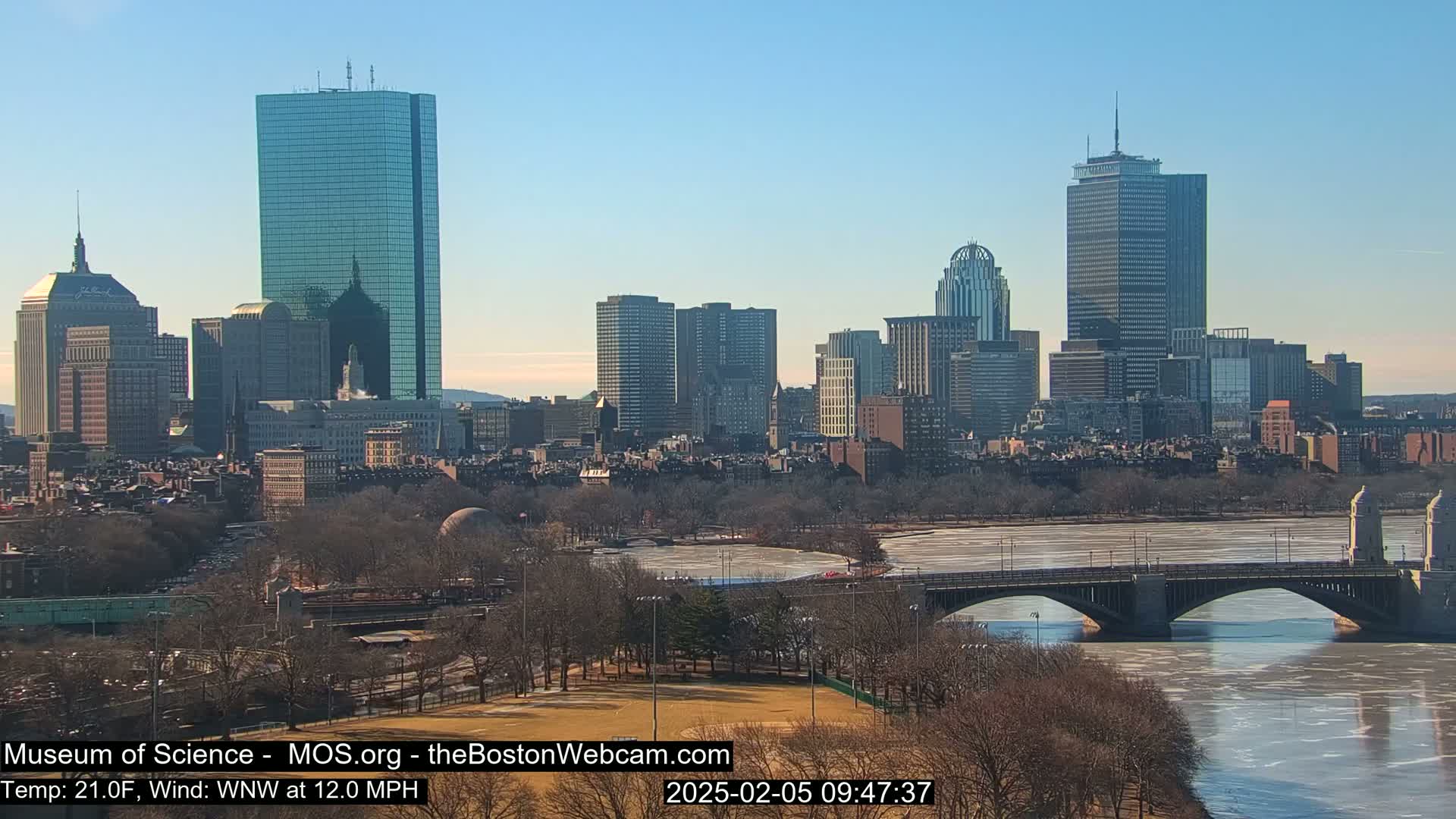 A city skyline with numerous skyscrapers is visible across a partly frozen river, under a clear blue sky.
