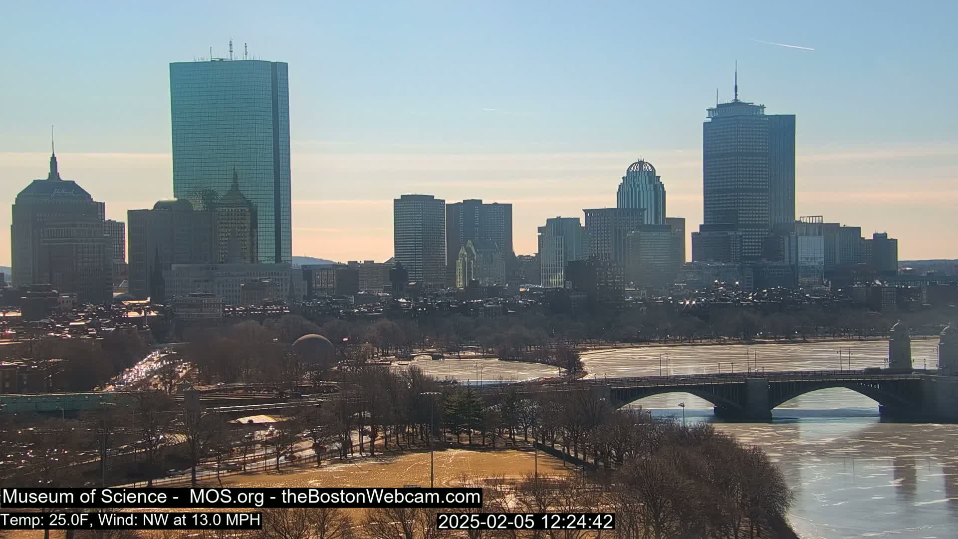 A sunny cityscape with several skyscrapers, a bridge over a partially frozen river, and bare winter trees under a clear sky.