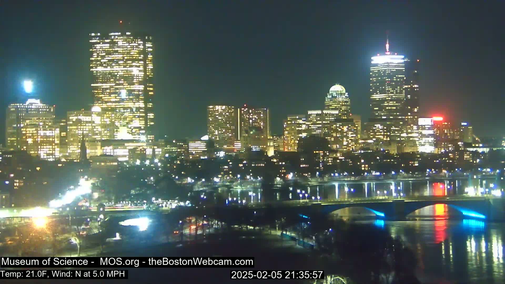 A nighttime cityscape with brightly lit skyscrapers and a bridge illuminated with blue lights across a dark river, under a clear sky with a north wind at 5 mph and a temperature of 21°F.