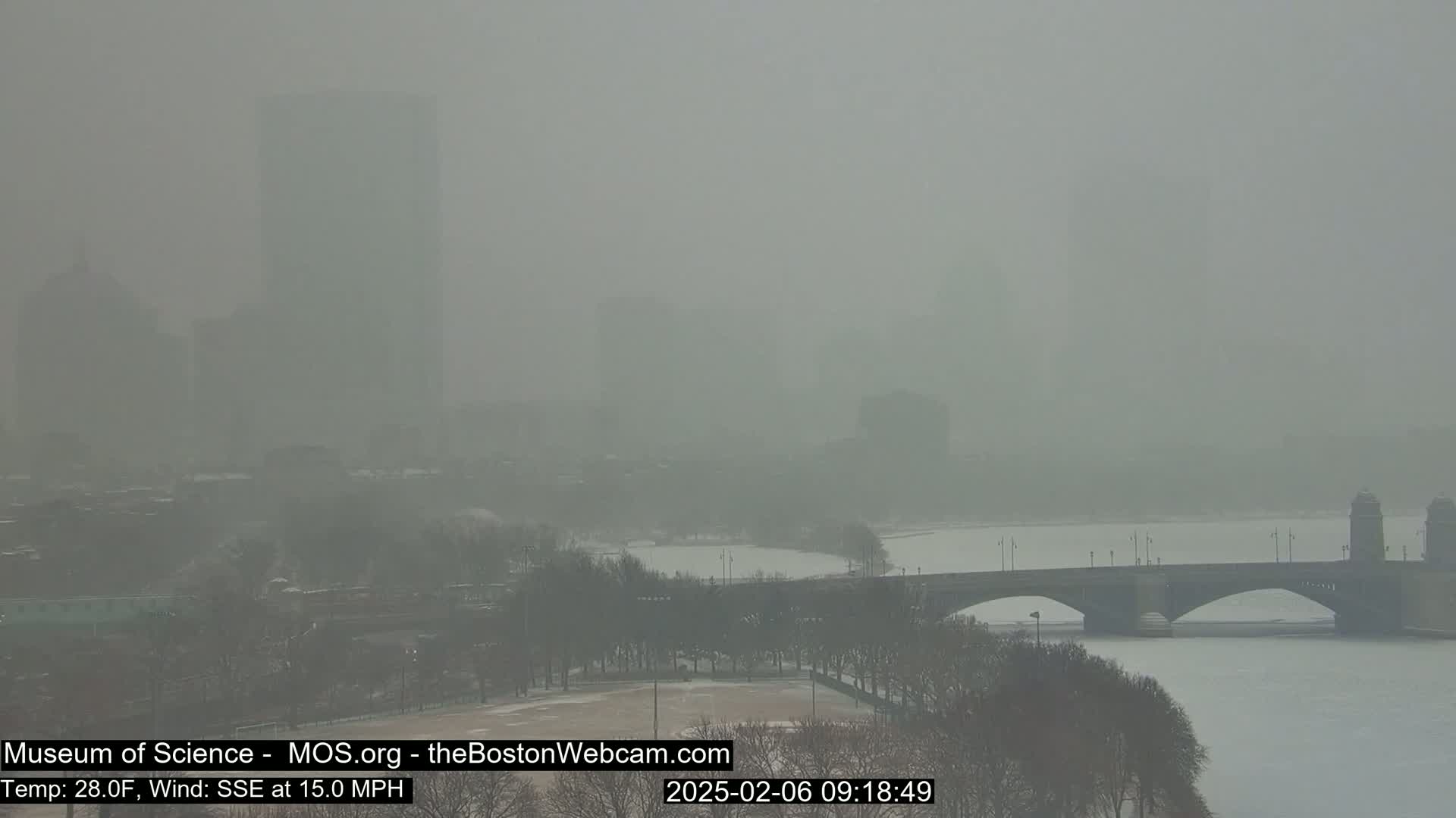 A hazy, snowy scene shows a bridge over a snow-covered river with buildings in the distance.