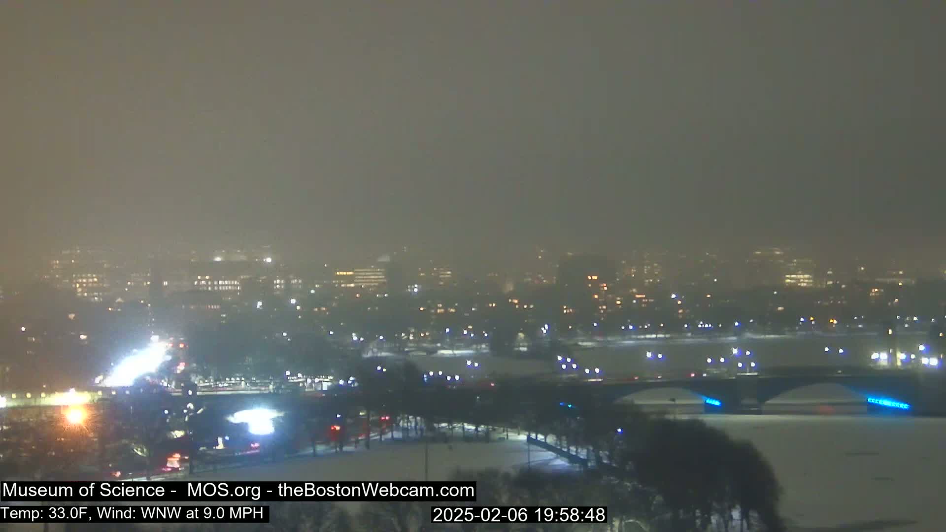 A snow-covered cityscape at night is viewed from a distance, with a bridge illuminated and vehicles visible on roads.