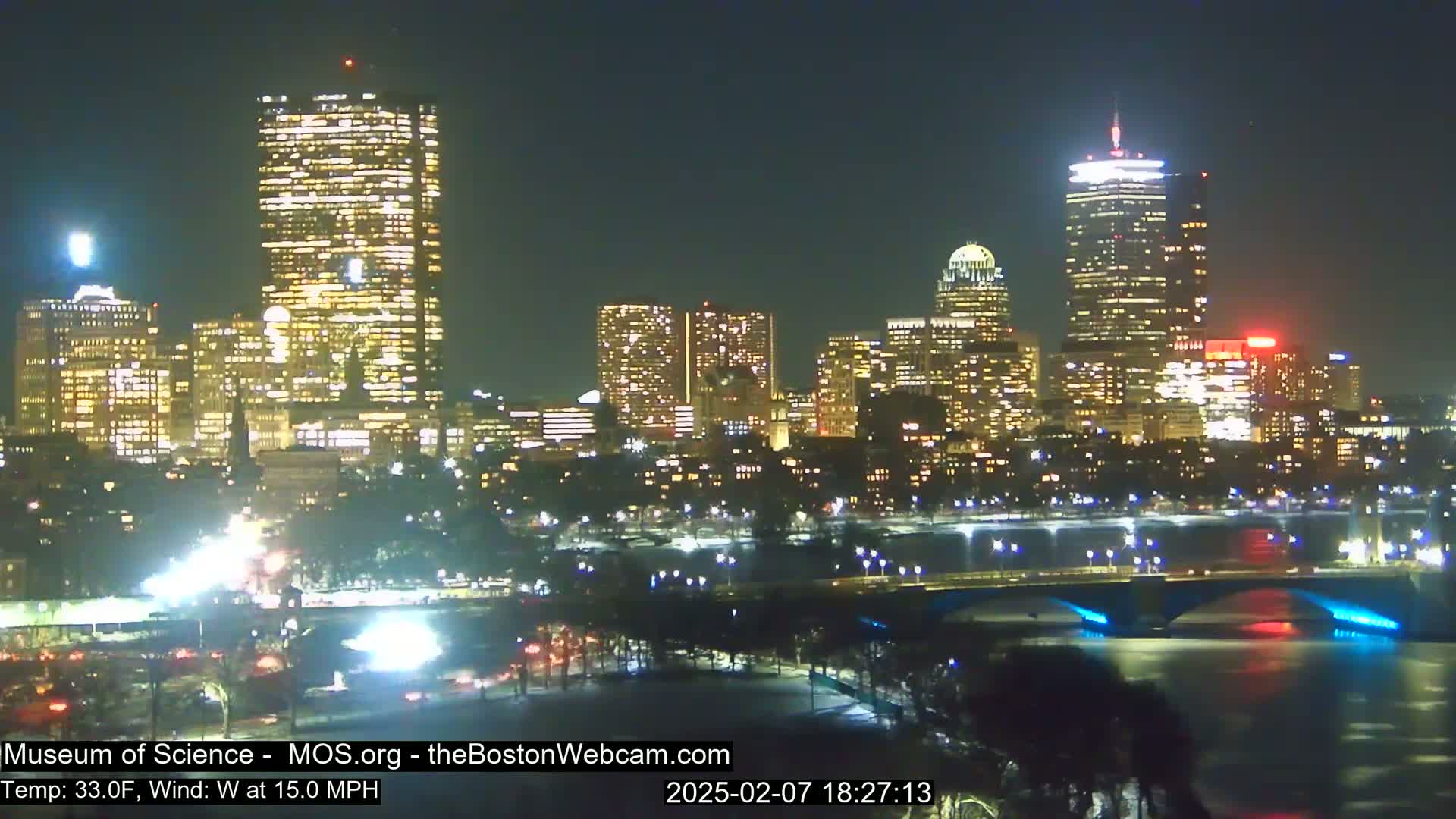 A nighttime view of a city skyline with illuminated buildings, a bridge, and a river, under a clear sky with a temperature of 33 degrees Fahrenheit and a 15 mph westerly wind.