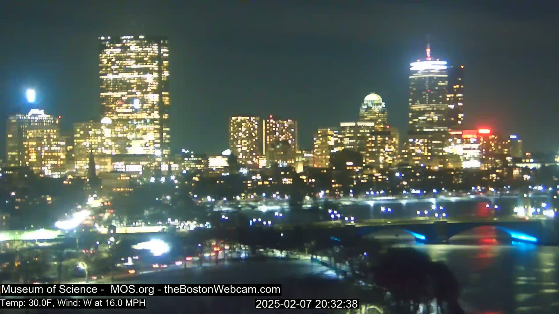 A nighttime cityscape with brightly lit buildings, a bridge, and a body of water under a clear sky, with a 30°F temperature and a 16 mph west wind.
