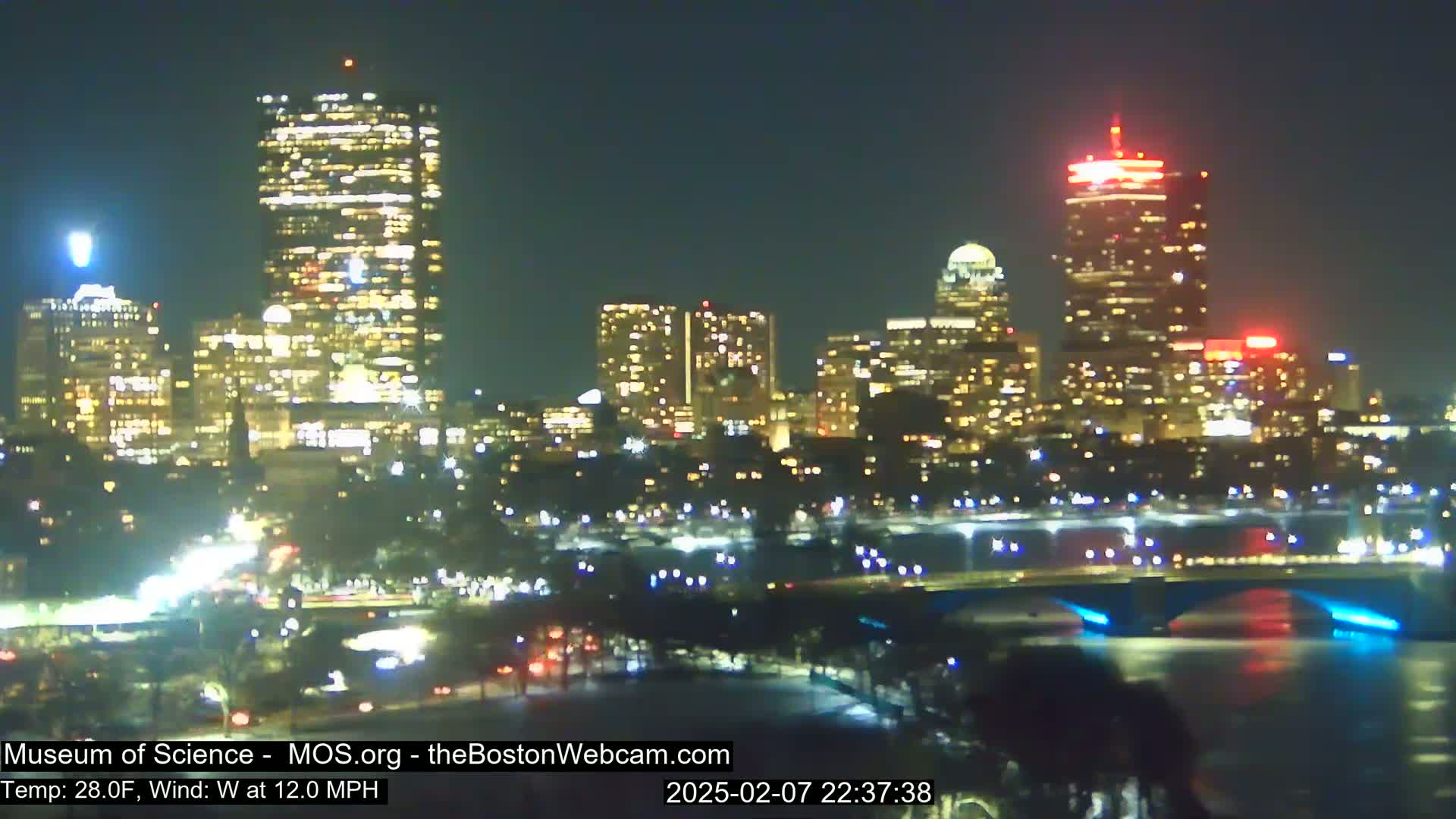 A nighttime view of a city skyline across a river, with a 28°F temperature and a 12 mph west wind.