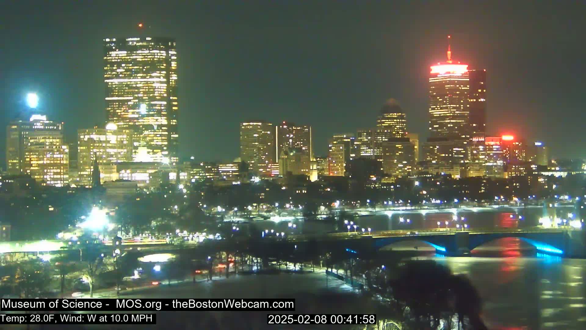 A nighttime cityscape with illuminated skyscrapers, bridges, and a river, under a clear sky with a 28°F temperature and a 10 mph west wind.